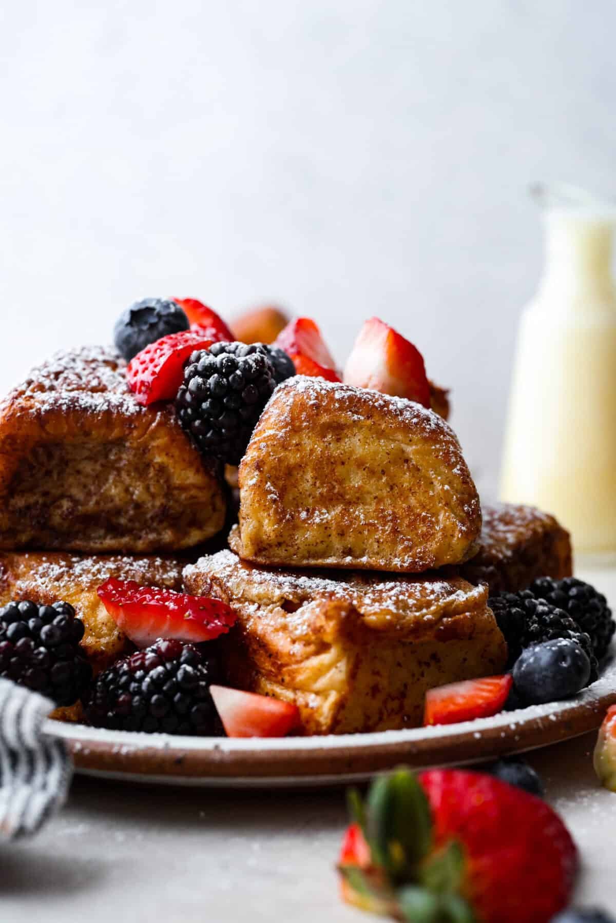 Hawaiian Roll French Toast is a fun twist on the classic, with sweet, buttery rolls that stay soft inside and turn golden outside. Instead of cutting them up, which made them too soggy, I used the whole rolls for the best texture. The real game changer was pairing them with my homemade coconut buttermilk syrup for an extra indulgent touch. Side shot of a pile of Hawaiian roll french toast with fresh berries and a dusting of powdered sugar.