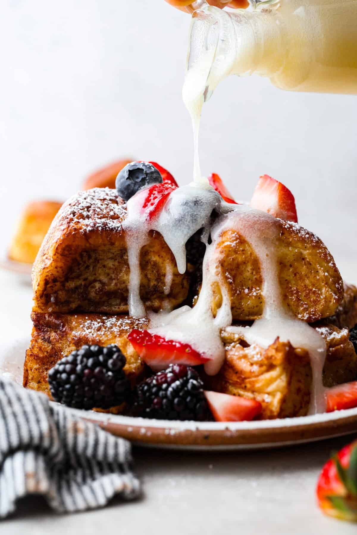 Hawaiian Roll French Toast is a fun twist on the classic, with sweet, buttery rolls that stay soft inside and turn golden outside. Instead of cutting them up, which made them too soggy, I used the whole rolls for the best texture. The real game changer was pairing them with my homemade coconut buttermilk syrup for an extra indulgent touch. Side shot of a pile of Hawaiian roll french toast with berries and someone pouring coconut buttermilk syrup over the top.