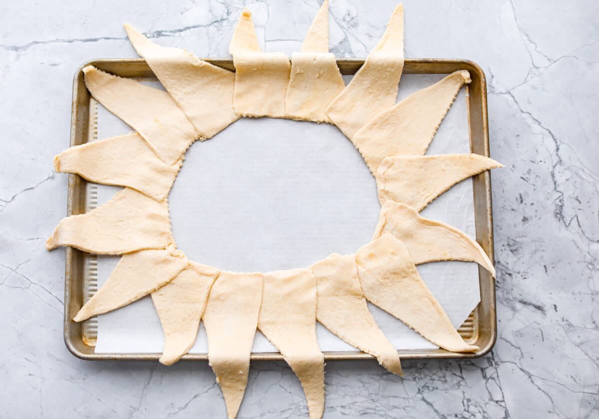 This Raspberry Cream Cheese Crescent Ring is a simple pastry with a flaky, golden crust, a sweet and tangy cream cheese filling, and a burst of raspberry flavor. Perfect for breakfast, brunch, or dessert, it's easy to make yet impressively delicious! Overhead shot of a baking sheet with parchment paper and crescent rolls spread out in a sunburst shape.