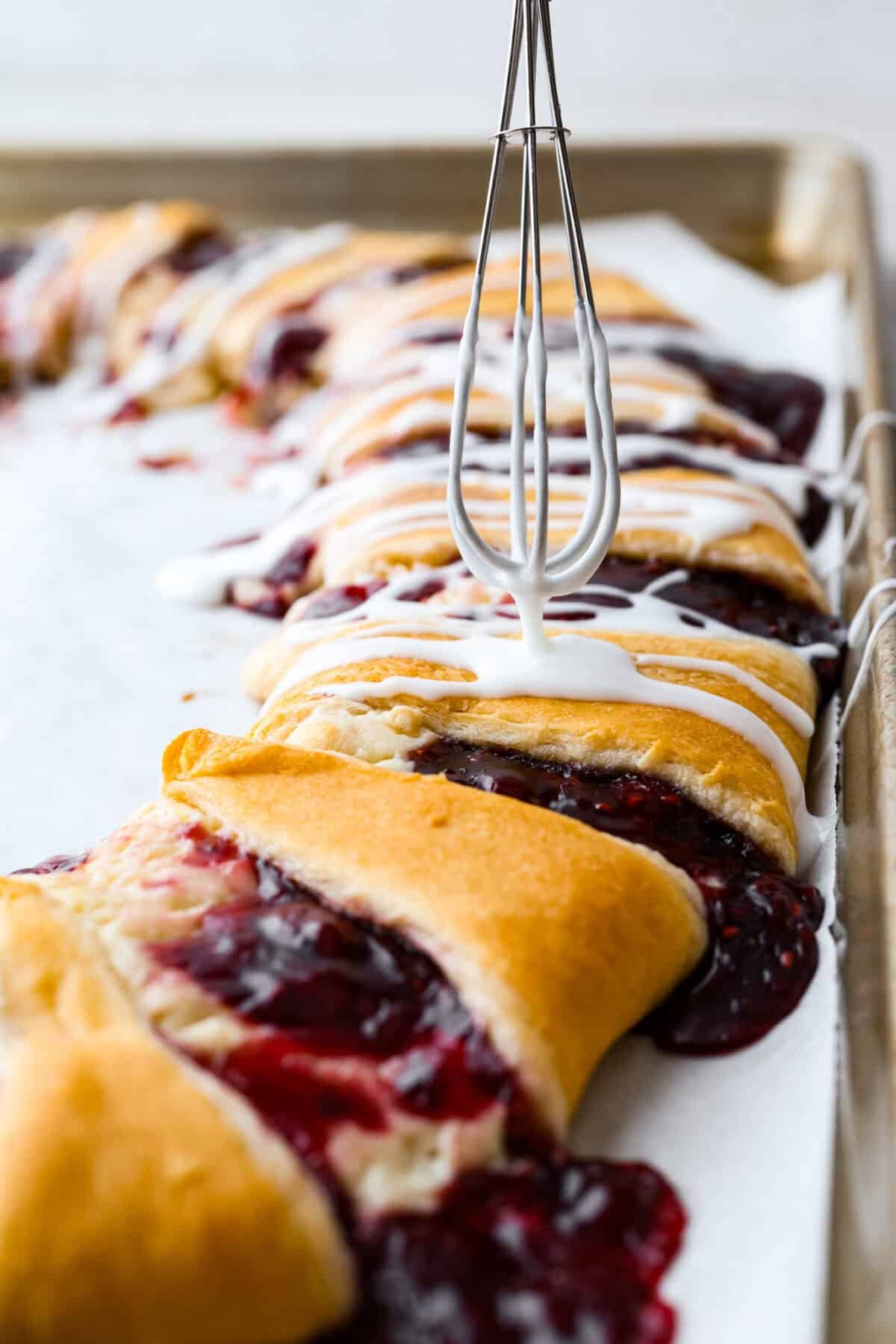 This Raspberry Cream Cheese Crescent Ring is a simple pastry with a flaky, golden crust, a sweet and tangy cream cheese filling, and a burst of raspberry flavor. Perfect for breakfast, brunch, or dessert, it's easy to make yet impressively delicious! Angle shot of someone drizzling glaze over the raspberry cream cheese crescent ring with a whisk.