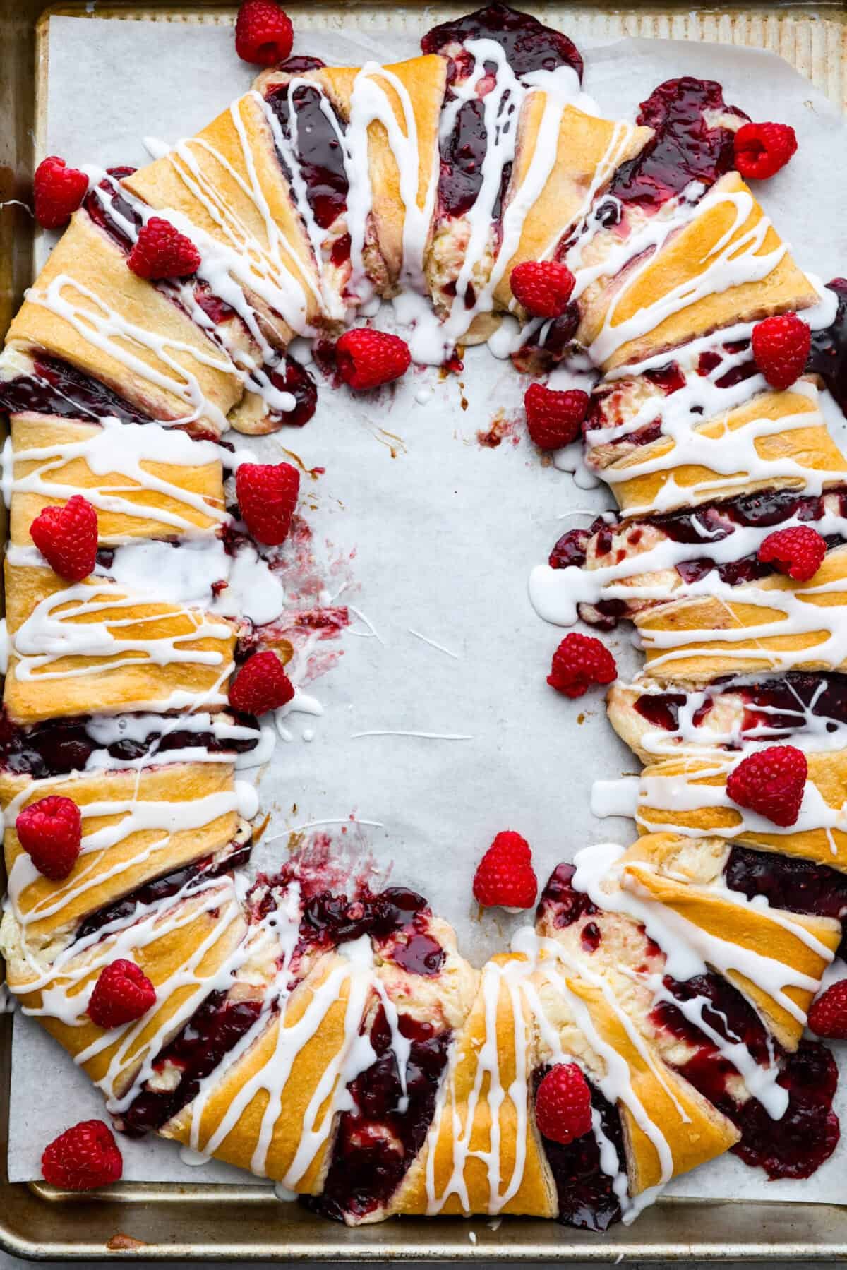 This Raspberry Cream Cheese Crescent Ring is a simple pastry with a flaky, golden crust, a sweet and tangy cream cheese filling, and a burst of raspberry flavor. Perfect for breakfast, brunch, or dessert, it's easy to make yet impressively delicious! Overhead shot of the baked raspberry cream cheese crescent roll ring with whole fresh raspberries sprinkled around.