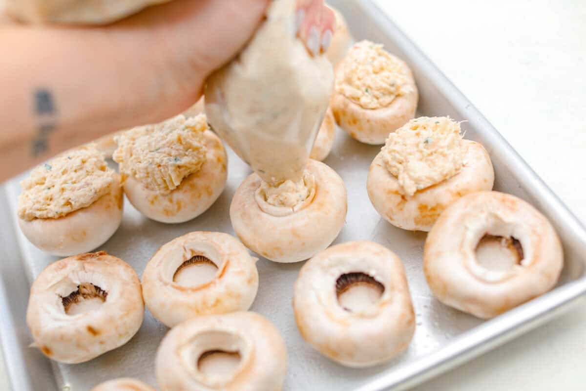These Crab Stuffed Mushrooms are a deliciously savory appetizer, perfect for impressing guests at any gathering. With a rich crab filling in every bite, they’re sure to be a crowd favorite! Overhead shot of someone piping crab stuffed mushroom filling into the de-stemmed mushrooms.