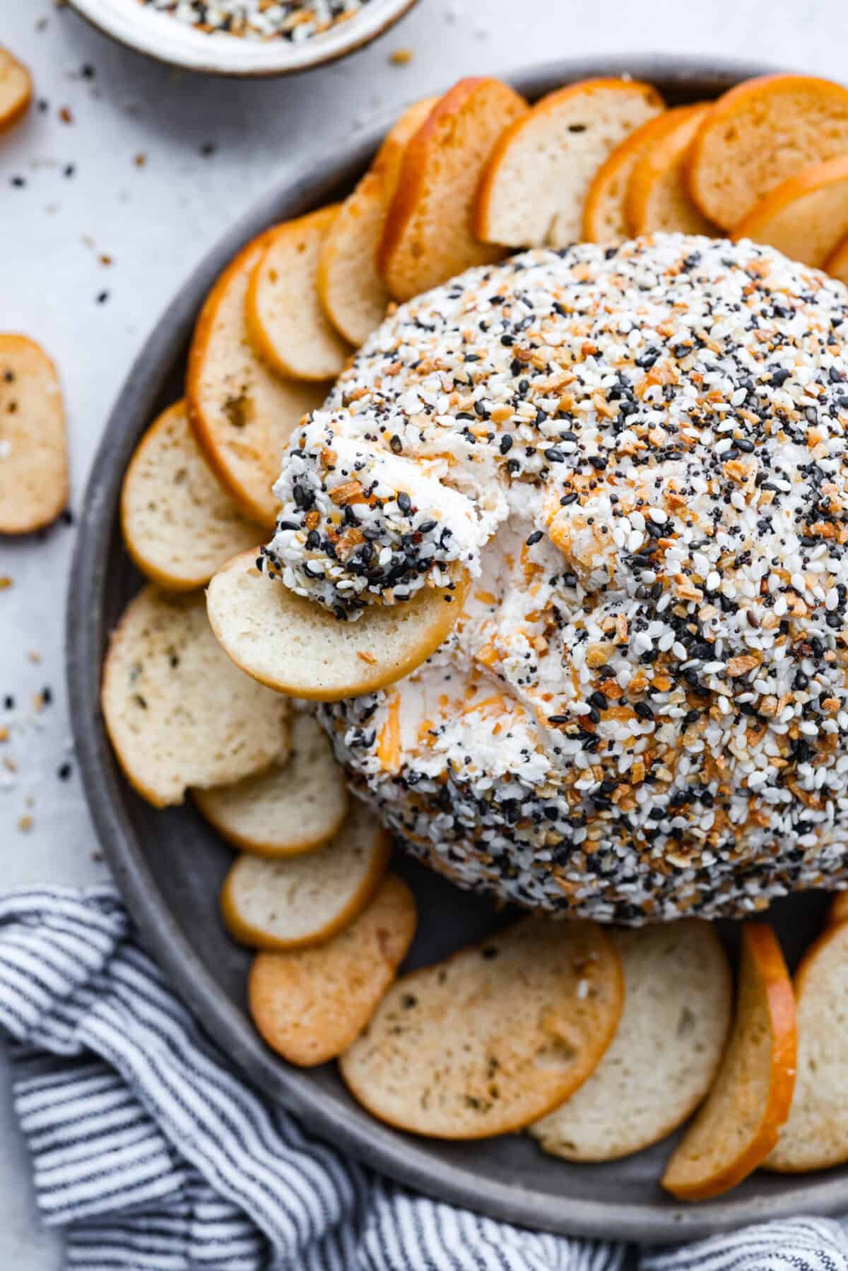 This everything bagel cheese ball combines the creamy richness of cheese with the savory blend of everything bagel seasoning! It's an irresistible appetizer you can serve with crackers, bagel chips, or veggies! Everyone will love it! Overhead shot of a crostini taking a scoop out of the cheese ball.