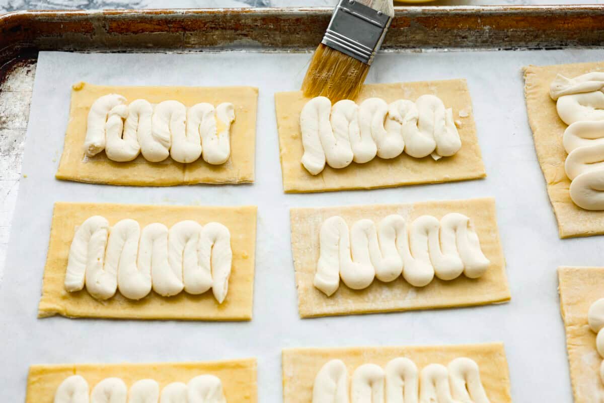 Craving the buttery goodness of a Starbucks cream cheese Danish? Now you can make it right at home! This easy copycat recipe uses flaky puff pastry and a sweet, creamy cheese filling for a treat that’s just as delicious, maybe even better! Overhead shot of someone brushing the egg and water mixture on to the exposed puff pastry around the filling.