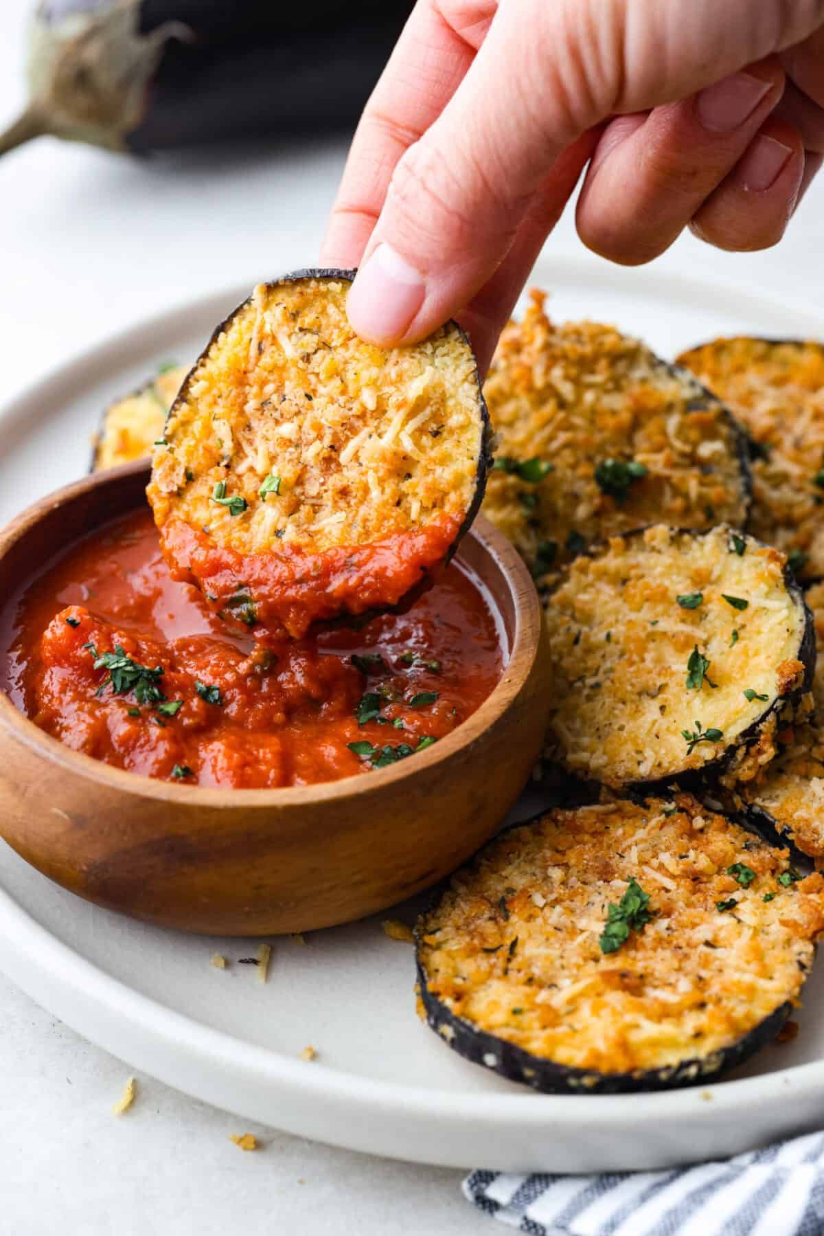 Crispy Air Fryer Eggplant is a fun flavor snack that is a healthier option than typical fried snacks. Tender eggplant slices are coated in crispy breadcrumbs and parmesan cheese, deliciously seasoned, then air-fried to perfection. Close up shot of someone dipping an ai fryer eggplant chip into marinara sauce.