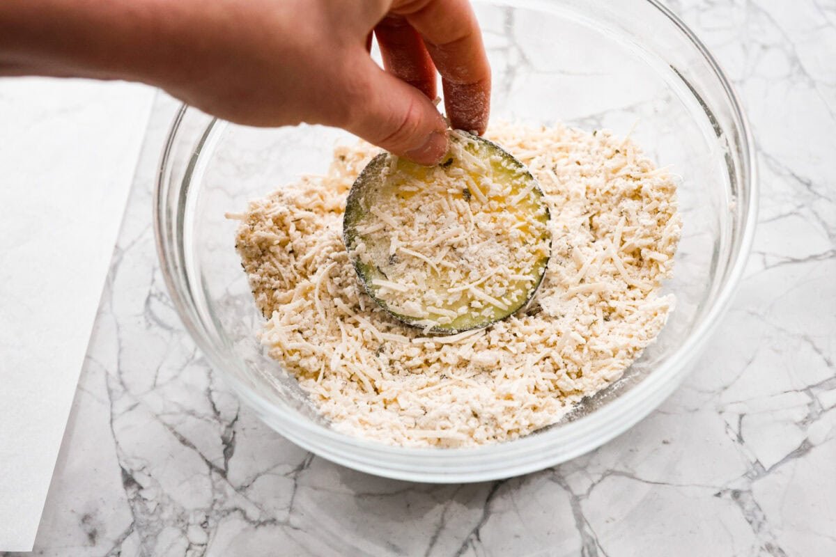 Crispy Air Fryer Eggplant is a fun flavor snack that is a healthier option than typical fried snacks. Tender eggplant slices are coated in crispy breadcrumbs and parmesan cheese, deliciously seasoned, then air-fried to perfection. Overhead shot of someone dipping a slice of egg washed eggplant into breading mixture.
