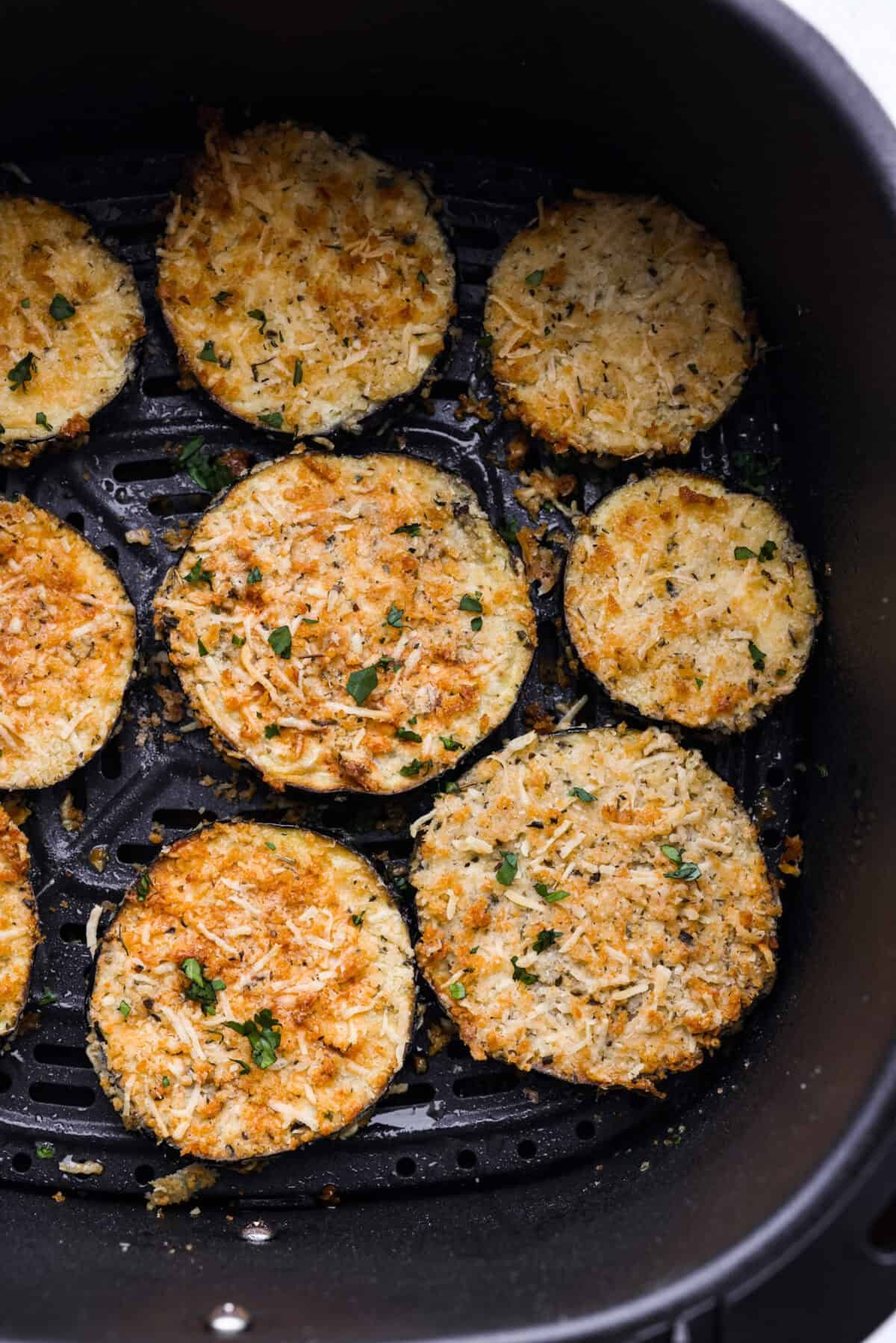 Crispy Air Fryer Eggplant is a fun flavor snack that is a healthier option than typical fried snacks. Tender eggplant slices are coated in crispy breadcrumbs and parmesan cheese, deliciously seasoned, then air-fried to perfection. Close up shot of air fried eggplant in the bottom of the air fryer basket.