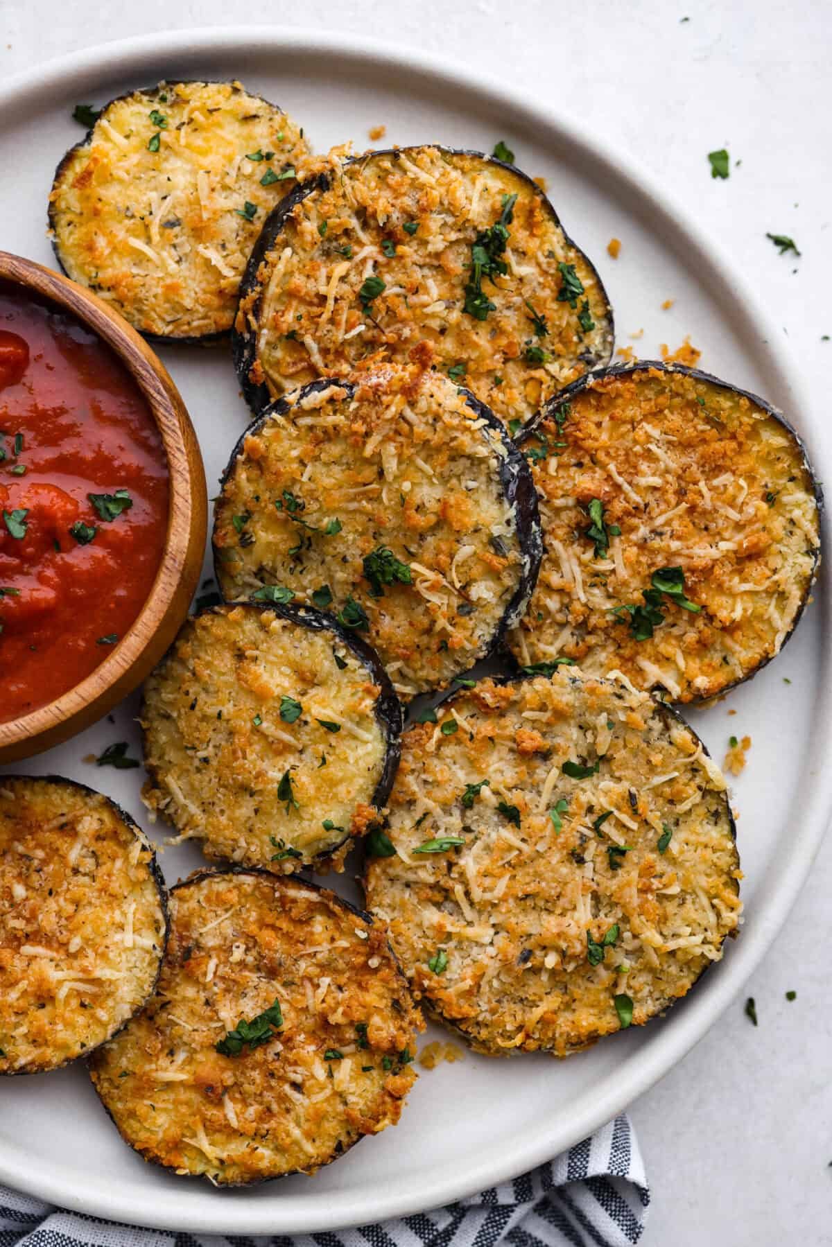 Crispy Air Fryer Eggplant is a fun flavor snack that is a healthier option than typical fried snacks. Tender eggplant slices are coated in crispy breadcrumbs and parmesan cheese, deliciously seasoned, then air-fried to perfection. Overhead shot of plated air fryer eggplant with a bowl of marinara sauce next to the slices.
