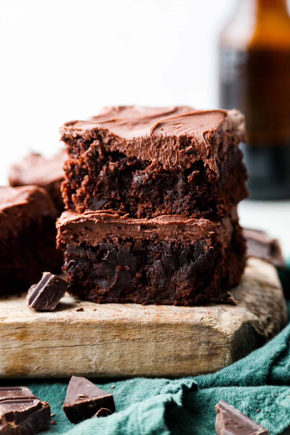 Rich, fudgy, and packed with deep chocolate flavor, Guinness Brownies are a must-try for any dessert lover! The stout beer enhances the cocoa, adding a subtle, malty depth of flavor that makes these brownies rich and irresistibly delicious. Two Guinness brownies stacked on top of each other on top of a wooden board with pieces of chocolate chunks scattered about.