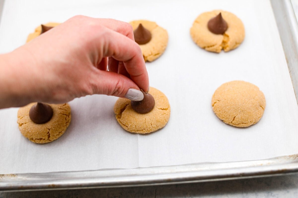 Peanut Butter Blossom Cookies are classic cookies that are perfect for every occasion. The delicious pairing of peanut butter and chocolate in these soft, buttery cookies will delight cookie lovers of all ages. Angle shot of someone placing a Hersheys Kiss in the center of a baked cookie dough ball.