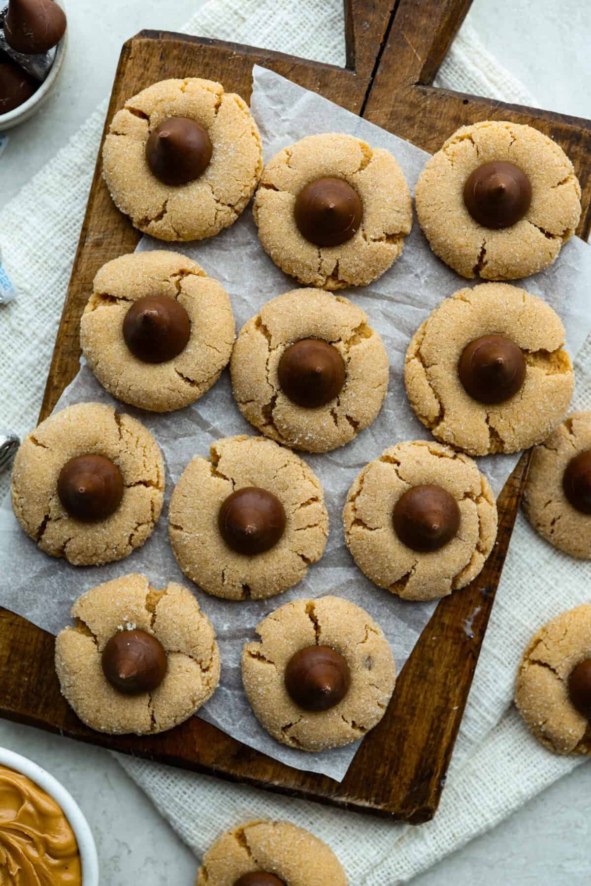 Peanut Butter Blossom Cookies are classic cookies that are perfect for every occasion. The delicious pairing of peanut butter and chocolate in these soft, buttery cookies will delight cookie lovers of all ages. Overhead shot of baked peanut butter blossom cookies on a wooden cutting board.