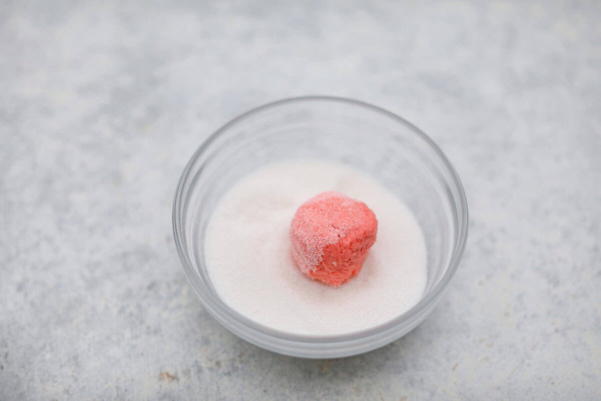 Strawberry Crinkle Cookies are the perfect blend of soft and chewy with a sweet, powdered sugar coating. Their vibrant color and classic strawberry cake flavor make them perfect for any occasion. Angle shot of a ball of dough rolled in granulated sugar.