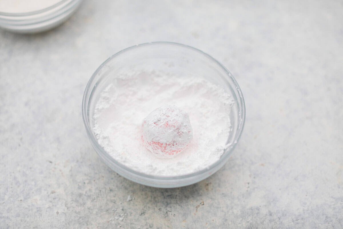Strawberry Crinkle Cookies are the perfect blend of soft and chewy with a sweet, powdered sugar coating. Their vibrant color and classic strawberry cake flavor make them perfect for any occasion. Angle shot of a ball of dough rolled in a bowl of powdered sugar.