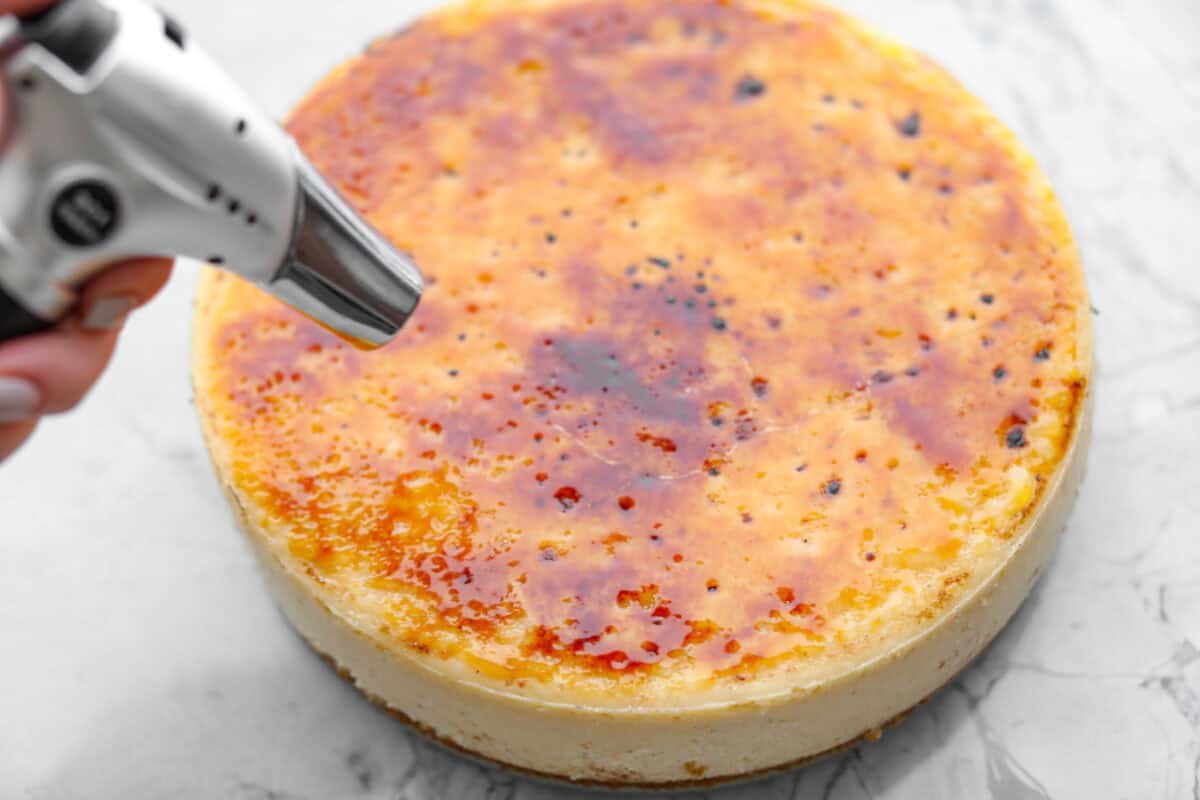 Creme Brulee Cheesecake is the perfect merging of two beloved desserts! It has the rich, creamy texture of cheesecake with the caramelized sugar topping of classic crème brûlée. This irresistible treat is the best of both worlds! Overhead shot of someone torching the sugar on top of the creme brûlée cheesecake.