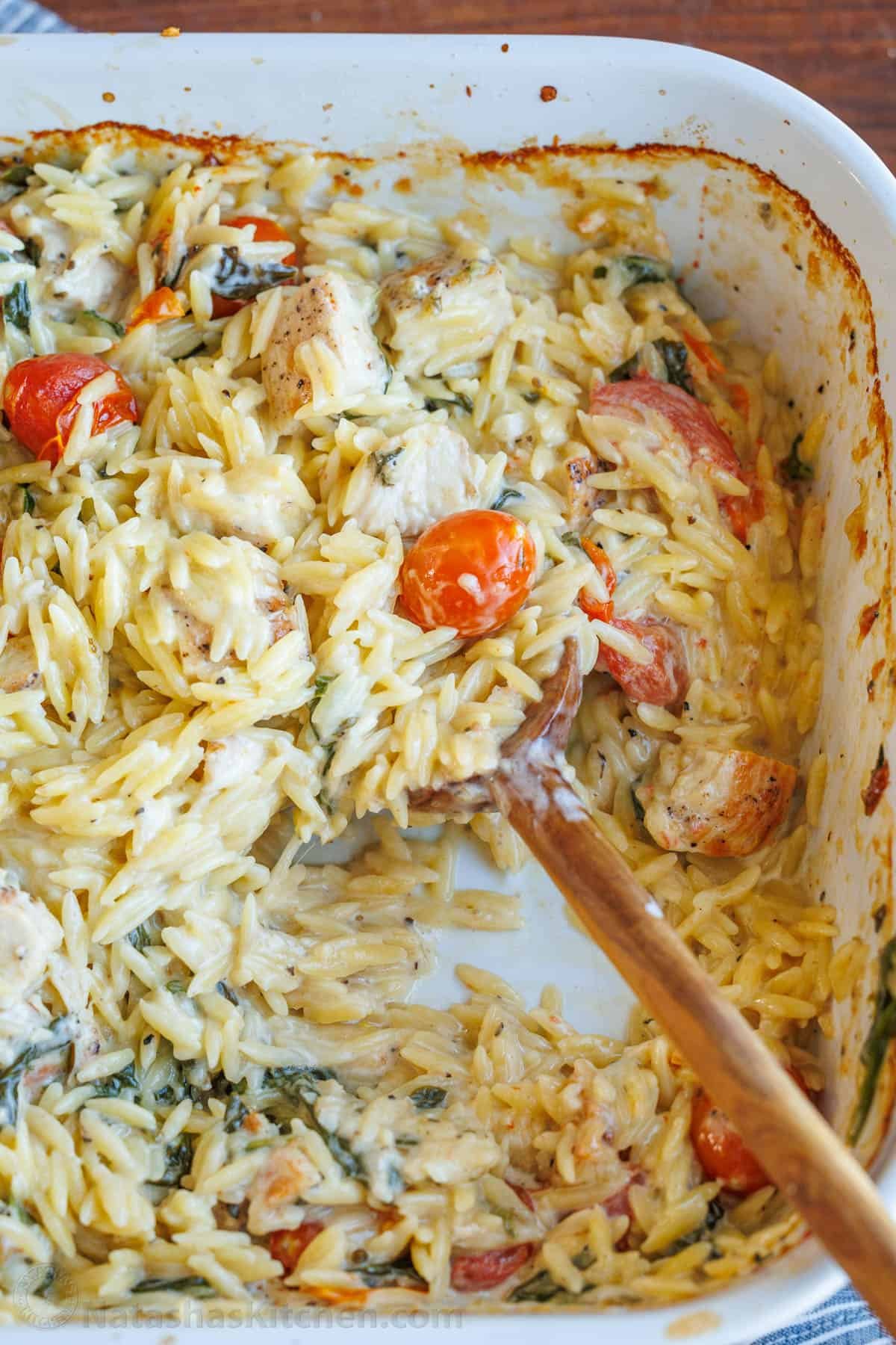 This Chicken Orzo Bake is an easy casserole that's creamy and cheesy. Mix it right in the dish, top it with cheese, and bake! Easy meal prep! A wooden spoon serving creamy chicken orzo bake casserole from a white dish