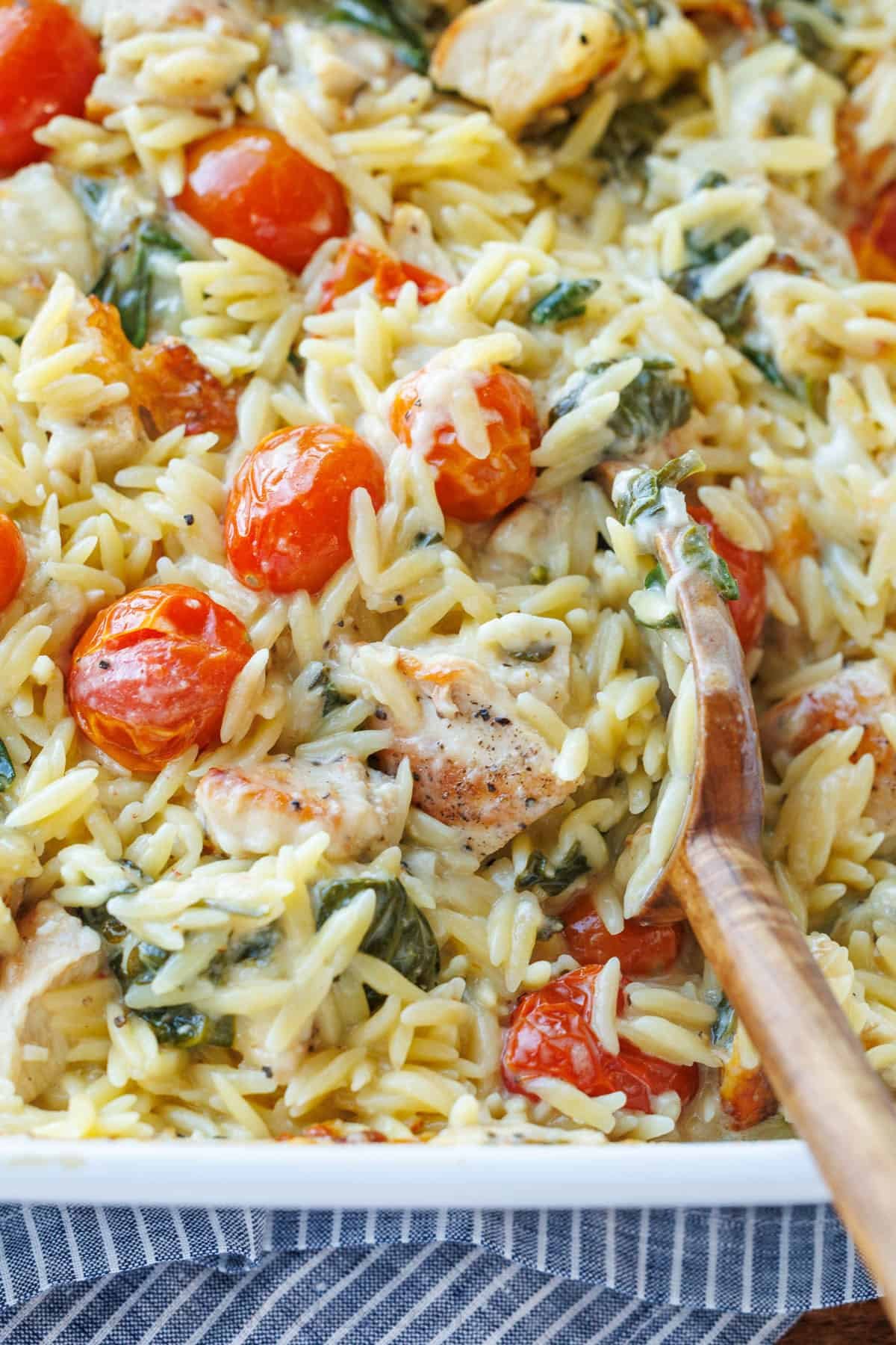 This Chicken Orzo Bake is an easy casserole that's creamy and cheesy. Mix it right in the dish, top it with cheese, and bake! Easy meal prep! A wooden spoon stirring creamy pasta casserole with tomatoes and spinach