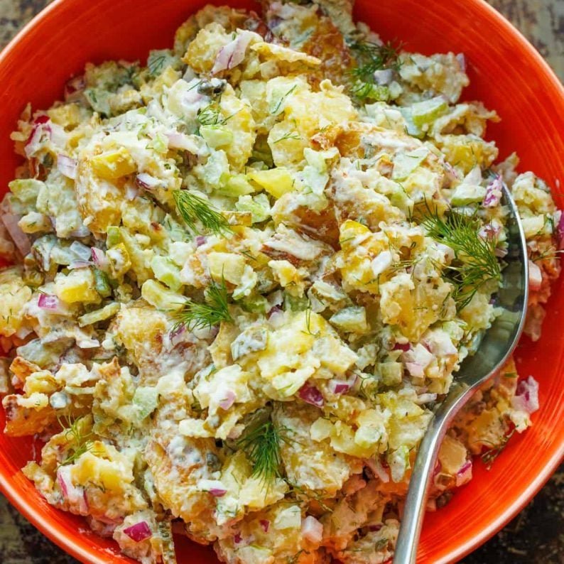 Smashed Potato Salad in a red serving bowl with a serving spoon, garnished with fresh dill.