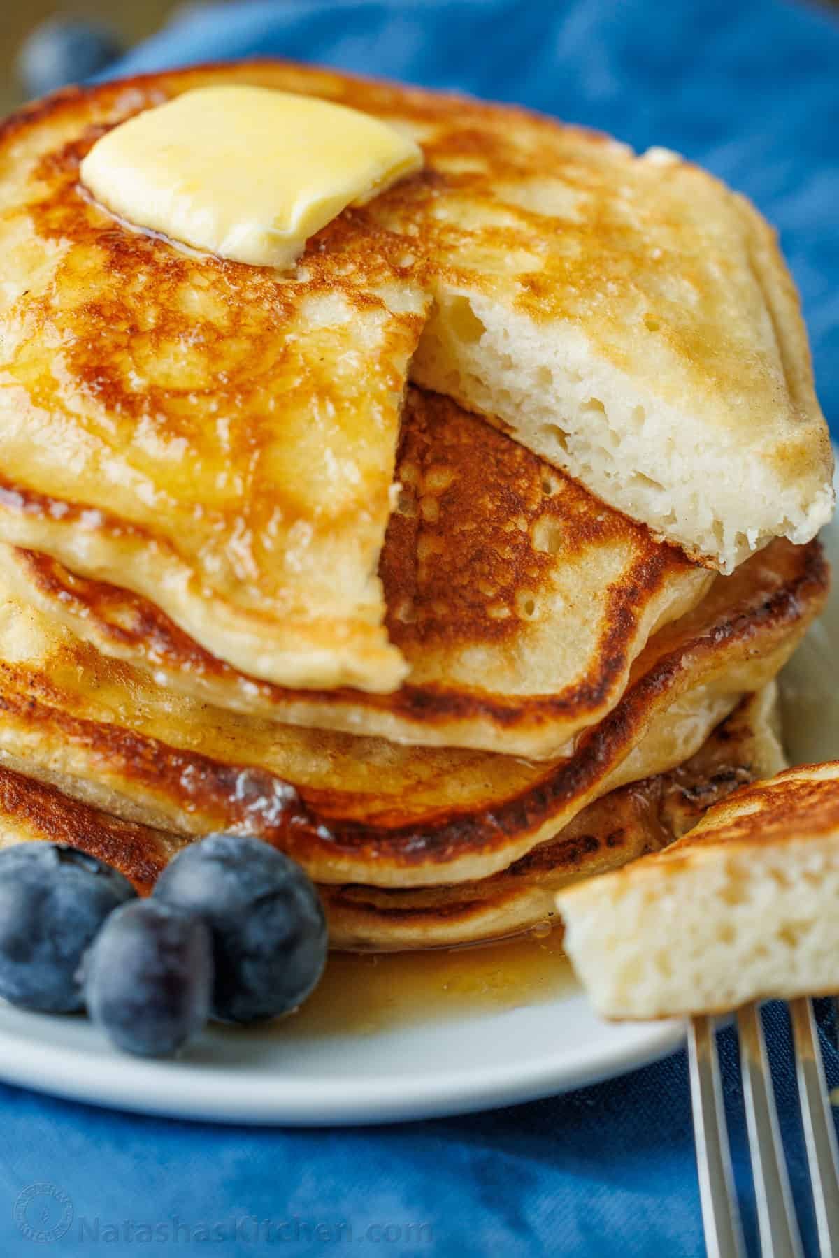 Overnight Sourdough Pancakes are easy! Mix everything before bed, leave at room temp overnight, and wake up to bubbly, ready-to-cook pancake batter. Sourdough Pancakes stacked on a plate with a pad of butter and maple syrup being poured over top. One bite has been sliced out of the top pancake. Blueberries garnish the plate.