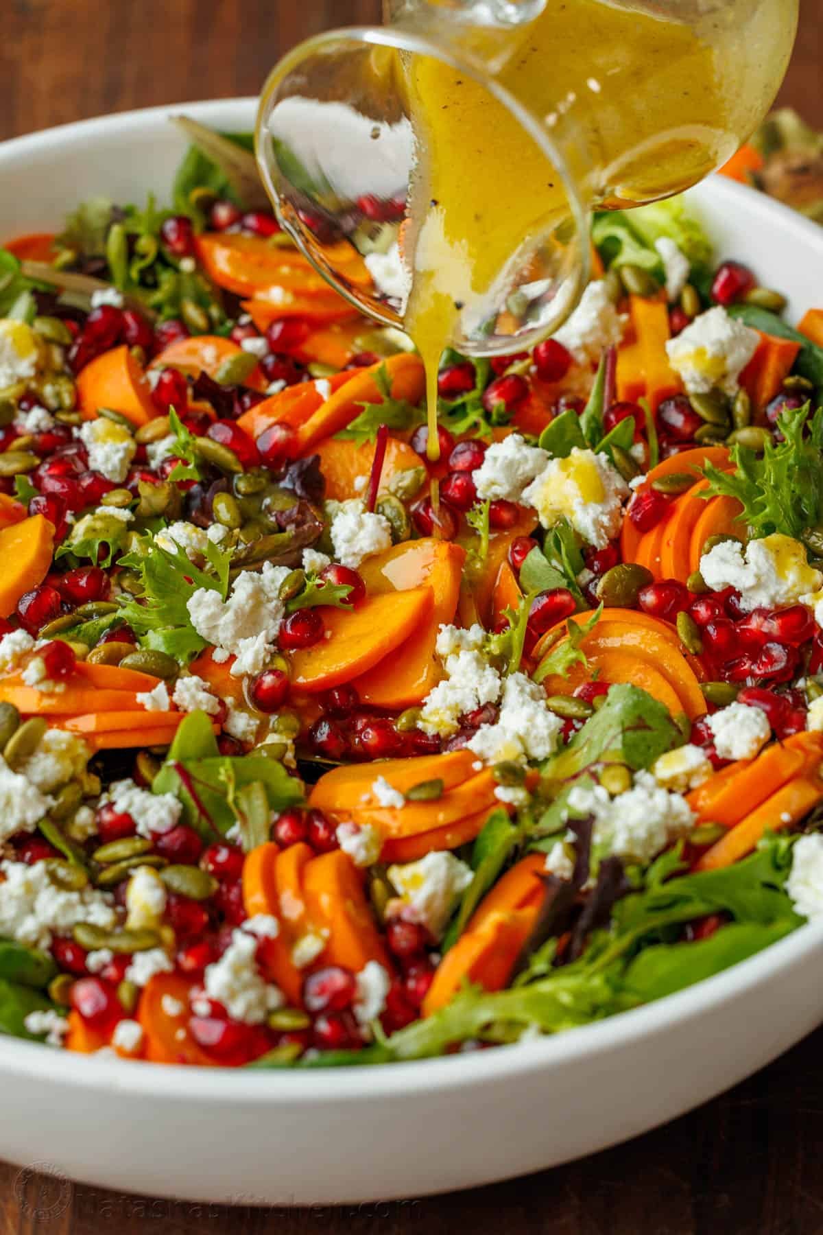 My persimmon Salad recipe features fresh fruit, crunchy toasted pepitas, tangy goat cheese, and a maple vinaigrette. Festive for any holiday! pouring dressing from a glass jar onto the persimmon salad