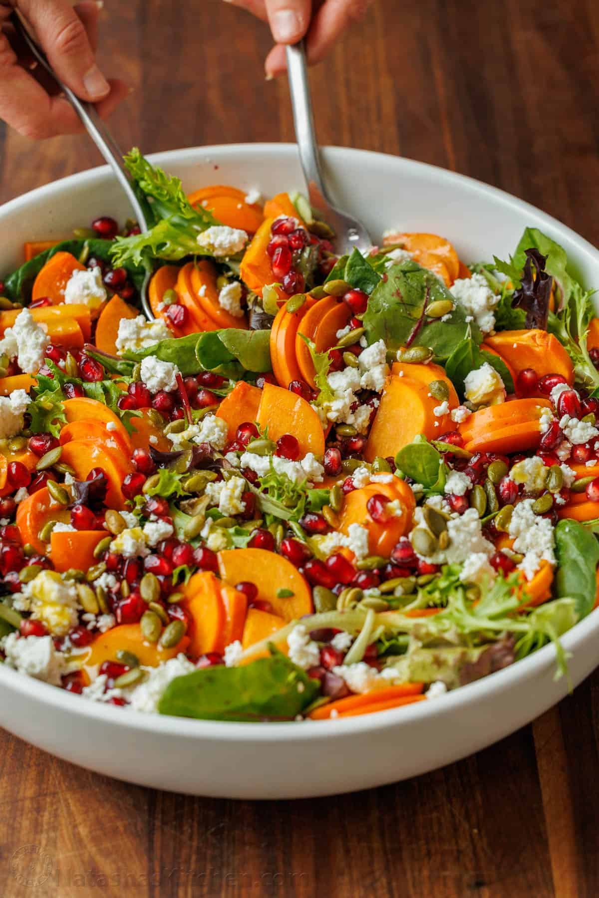 My persimmon Salad recipe features fresh fruit, crunchy toasted pepitas, tangy goat cheese, and a maple vinaigrette. Festive for any holiday! Serving persimmon salad with utensils out of a shite bowl