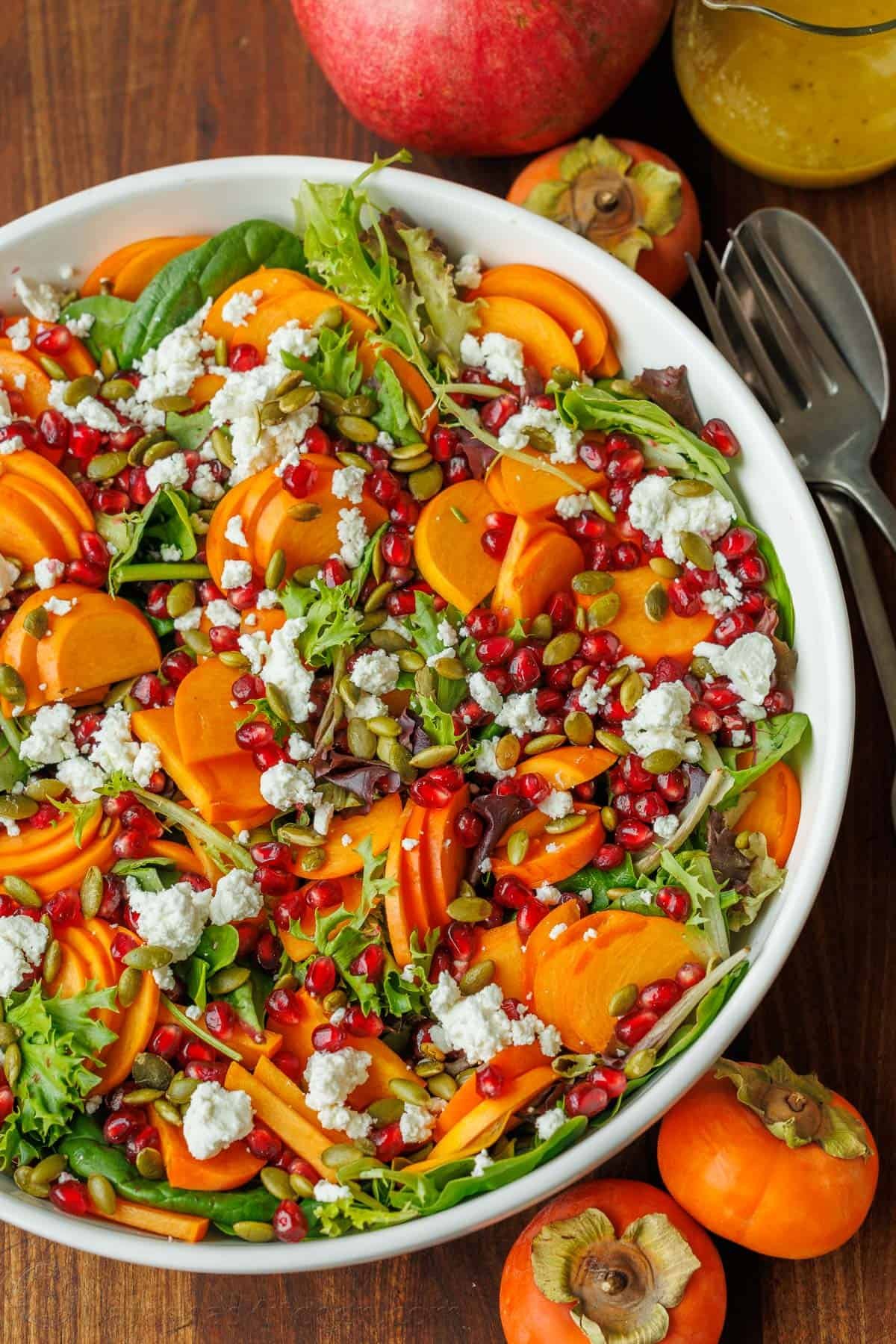 My persimmon Salad recipe features fresh fruit, crunchy toasted pepitas, tangy goat cheese, and a maple vinaigrette. Festive for any holiday! Holiday side dish in a white bowl with utensils and fresh winter fruit
