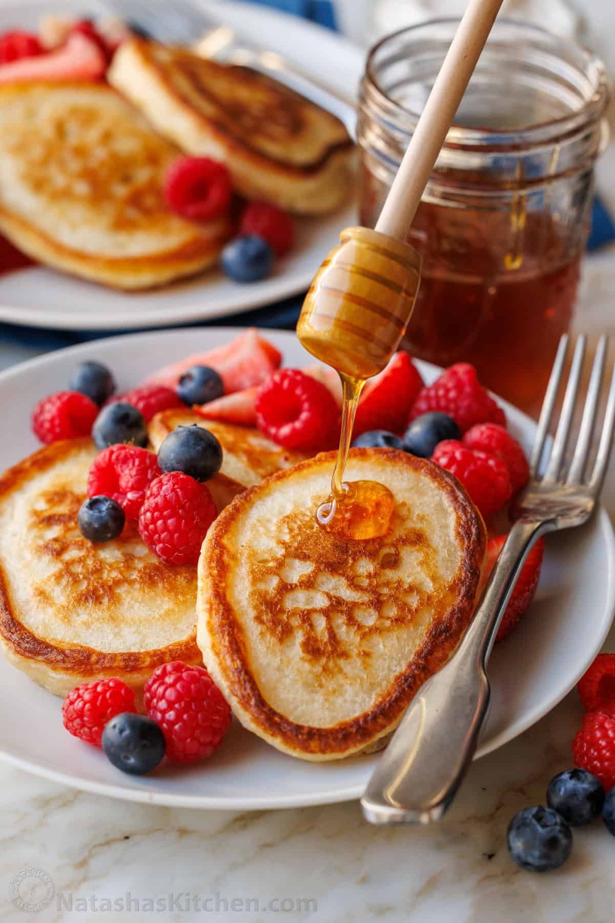 Sourdough Discard Pancakes are a delightful twist on the classic breakfast favorite, with a soft and spongy texture infused with tangy flavor from sourdough starter. They're a satisfying treat! Sourdough discard pancakes with honey being drizzled on top with a honey wand. Mixed berries are scattered on top of the pancakes on a white plate.
