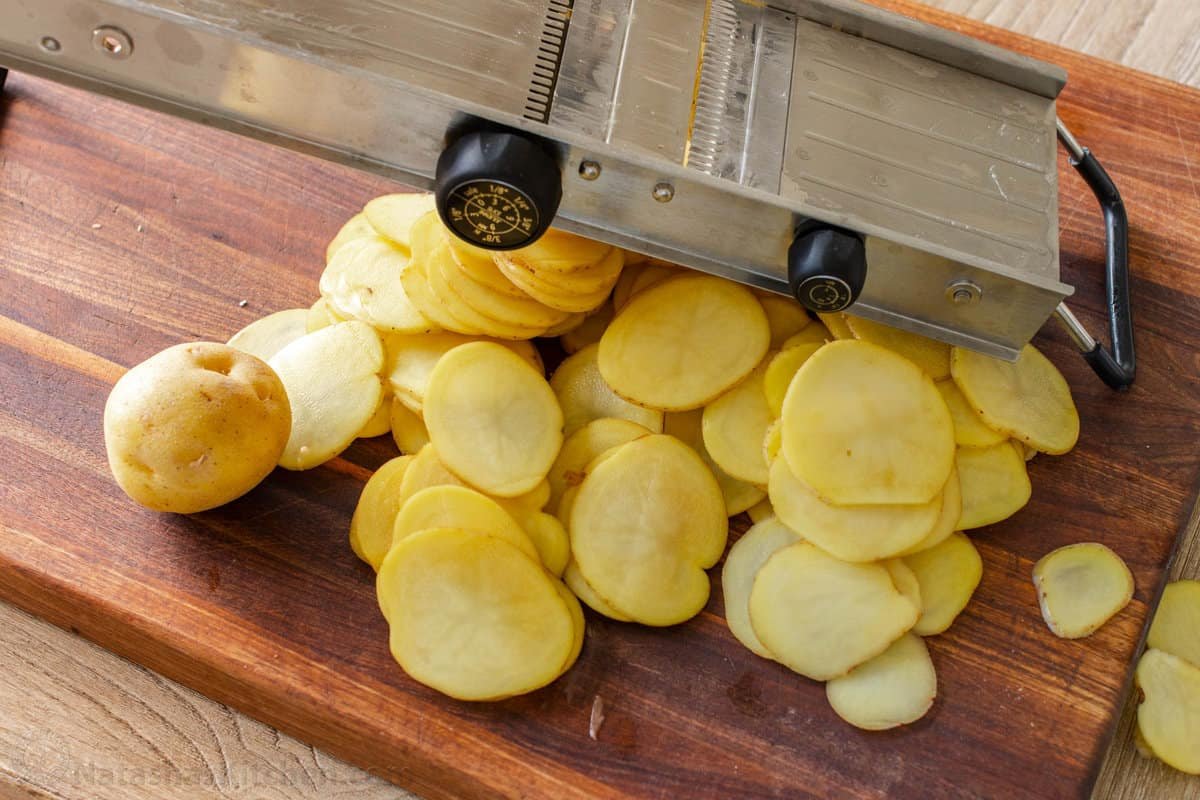 Creamy Au Gratin Potatoes are an easy holiday side dish, made with thinly sliced potatoes baked in a rich and velvety cheese sauce. Thin potato slices on a wooden cutting board below a mandolin.