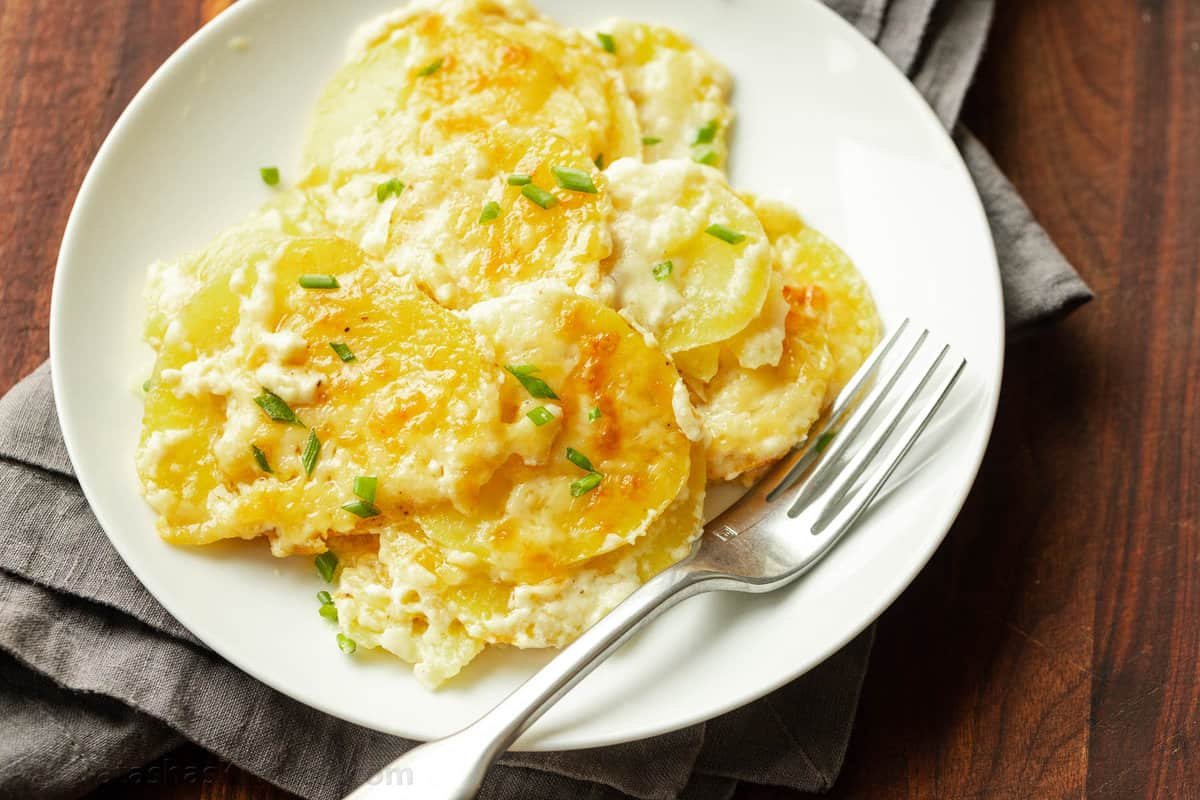 Creamy Au Gratin Potatoes are an easy holiday side dish, made with thinly sliced potatoes baked in a rich and velvety cheese sauce. Au gratin potatoes on a white plate with a fork.