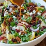 Pear Salad being dressed with Balsamic Vinaigrette in a white bowl, loaded with a variety of pears, red onion, goat cheese, cranberries, and toasted pecans