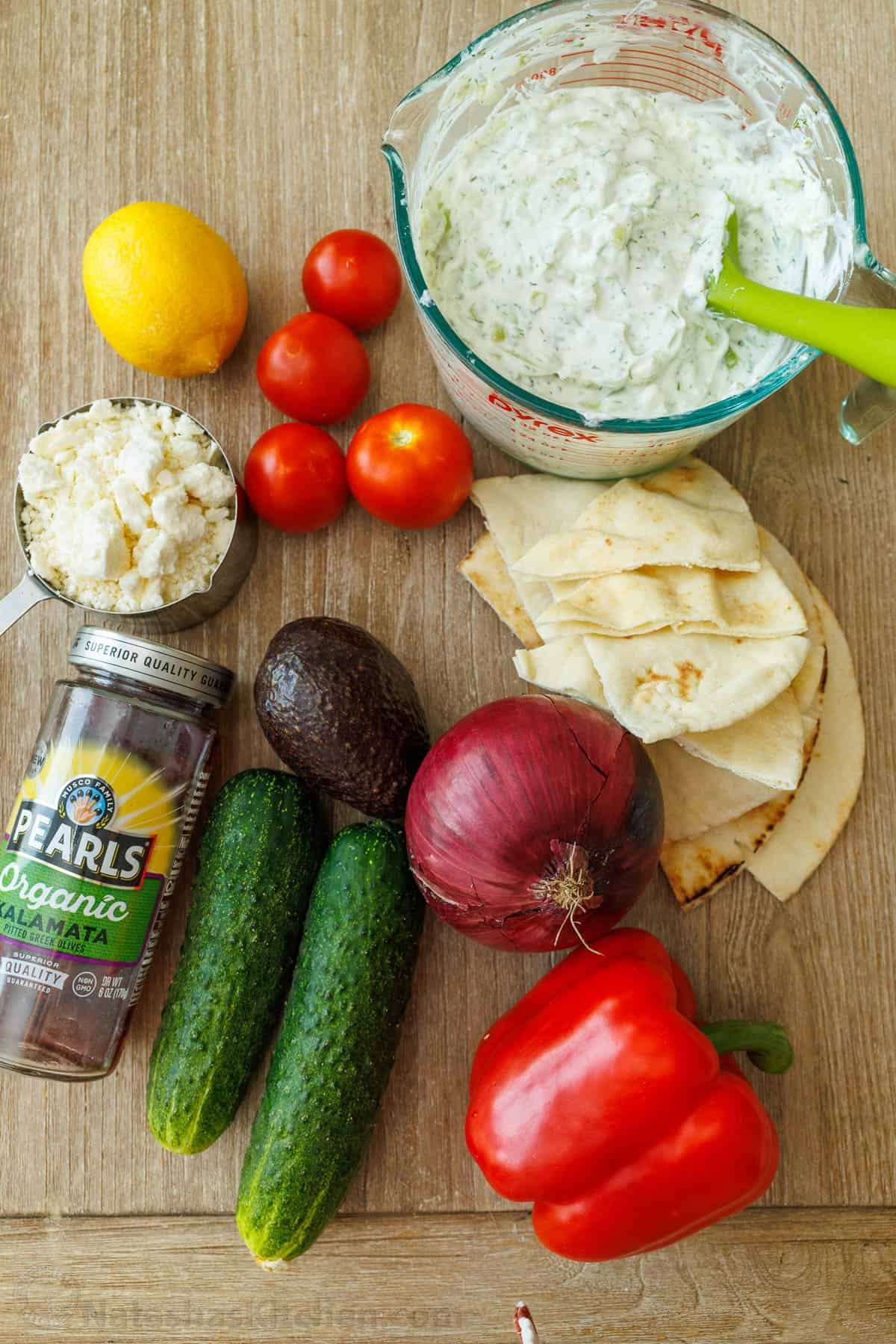 Greek Chicken Bowls salad ingredients: tzatziki sauce, bell pepper, tomatoes, avocado, cucumber, red onion, kalamata olives, feta cheese, lemon wedges