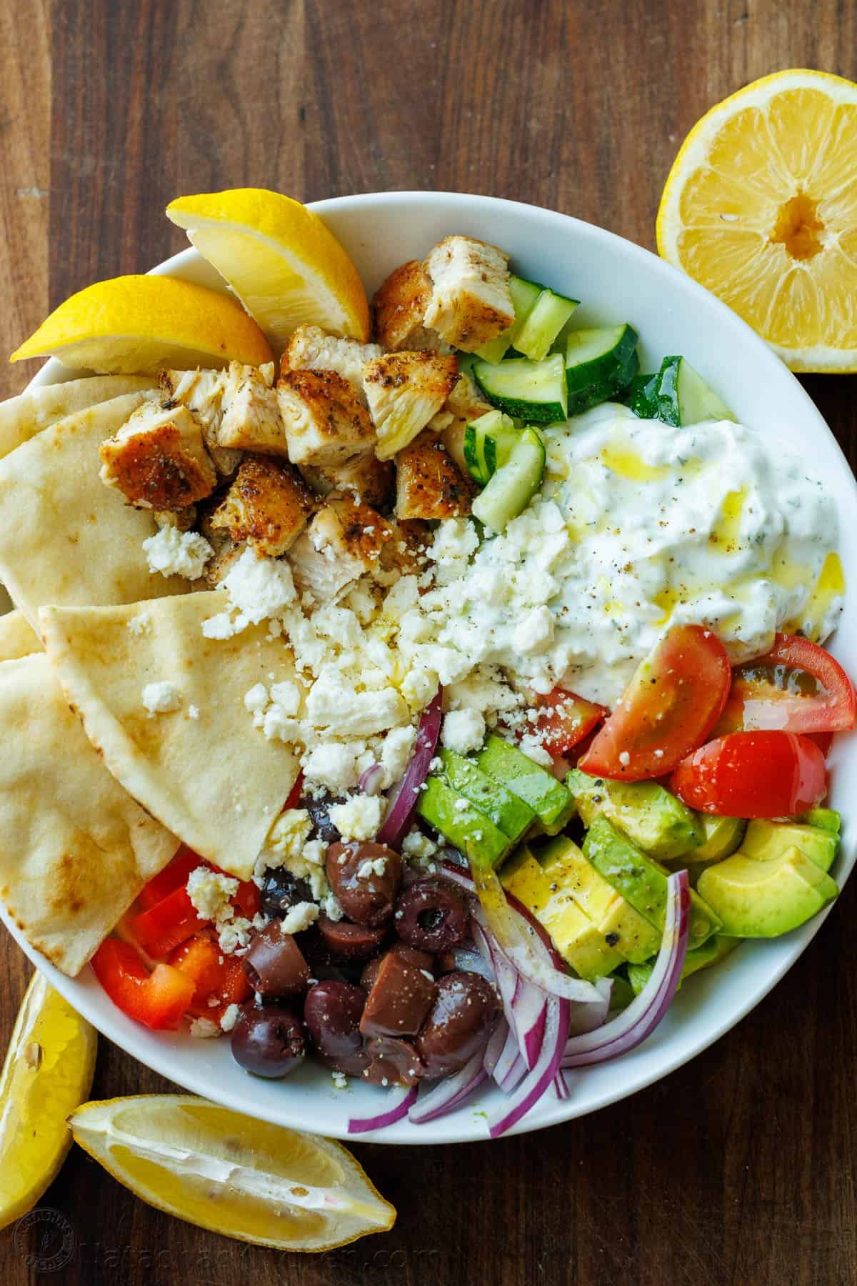 Greek Chicken Bowls combine the classic elements of a Greek Salad—cucumbers, tomatoes, olives, and feta—elevated with juicy grilled chicken and a refreshing dollop of tzatziki. Chopped Greek Chicken Bowl ingredients in a white bowl with lemon wedges and pita bread
