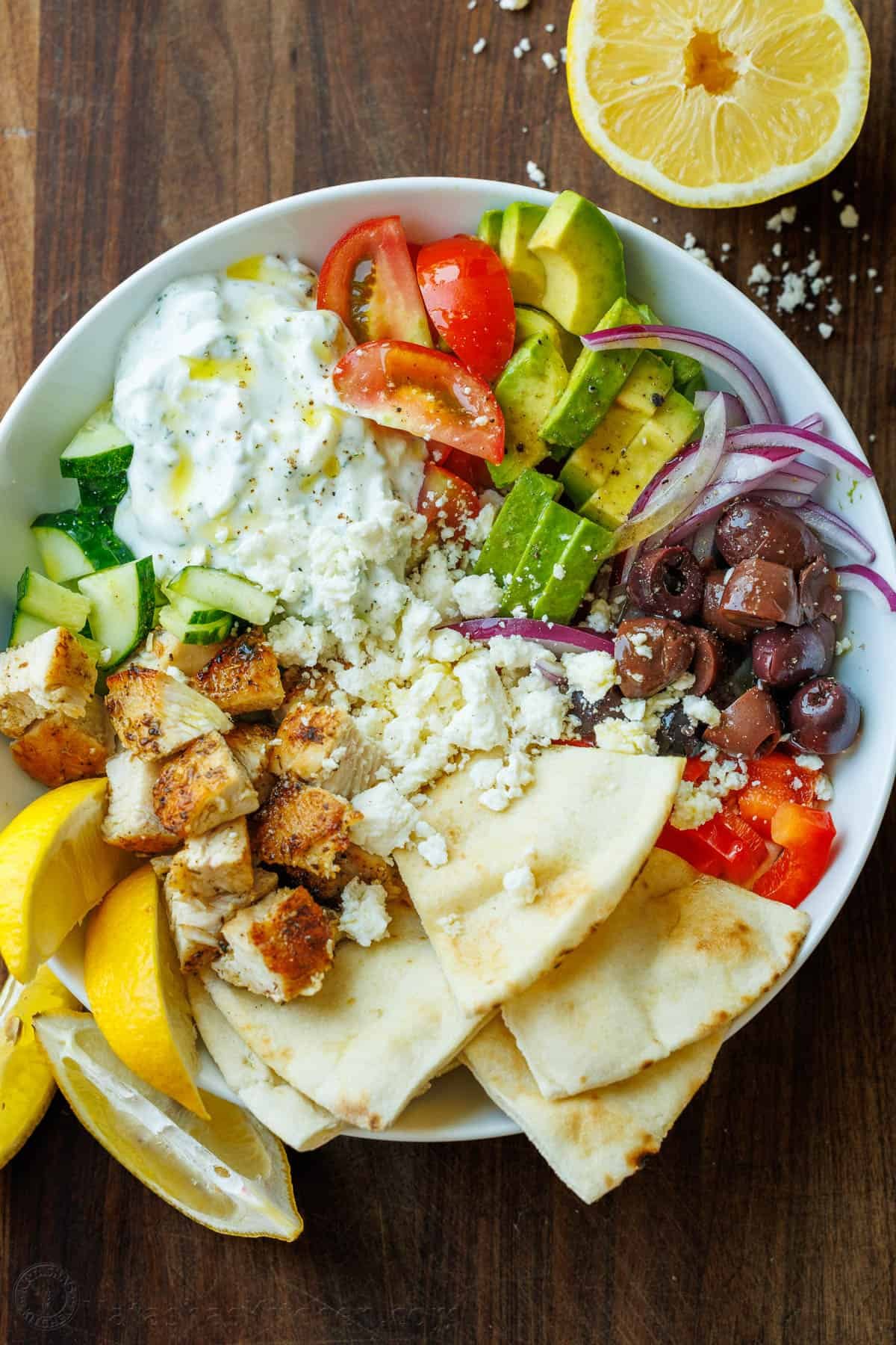 Greek Chicken Bowls combine the classic elements of a Greek Salad—cucumbers, tomatoes, olives, and feta—elevated with juicy grilled chicken and a refreshing dollop of tzatziki. Chopped Greek Chicken Bowl ingredients in a white bowl with lemon wedges and pita bread