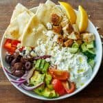 Greek Chicken Bowl with chopped ingredients, tzatziki sauce, homemade pita bread, and lemon wedges, served in a white bowl.