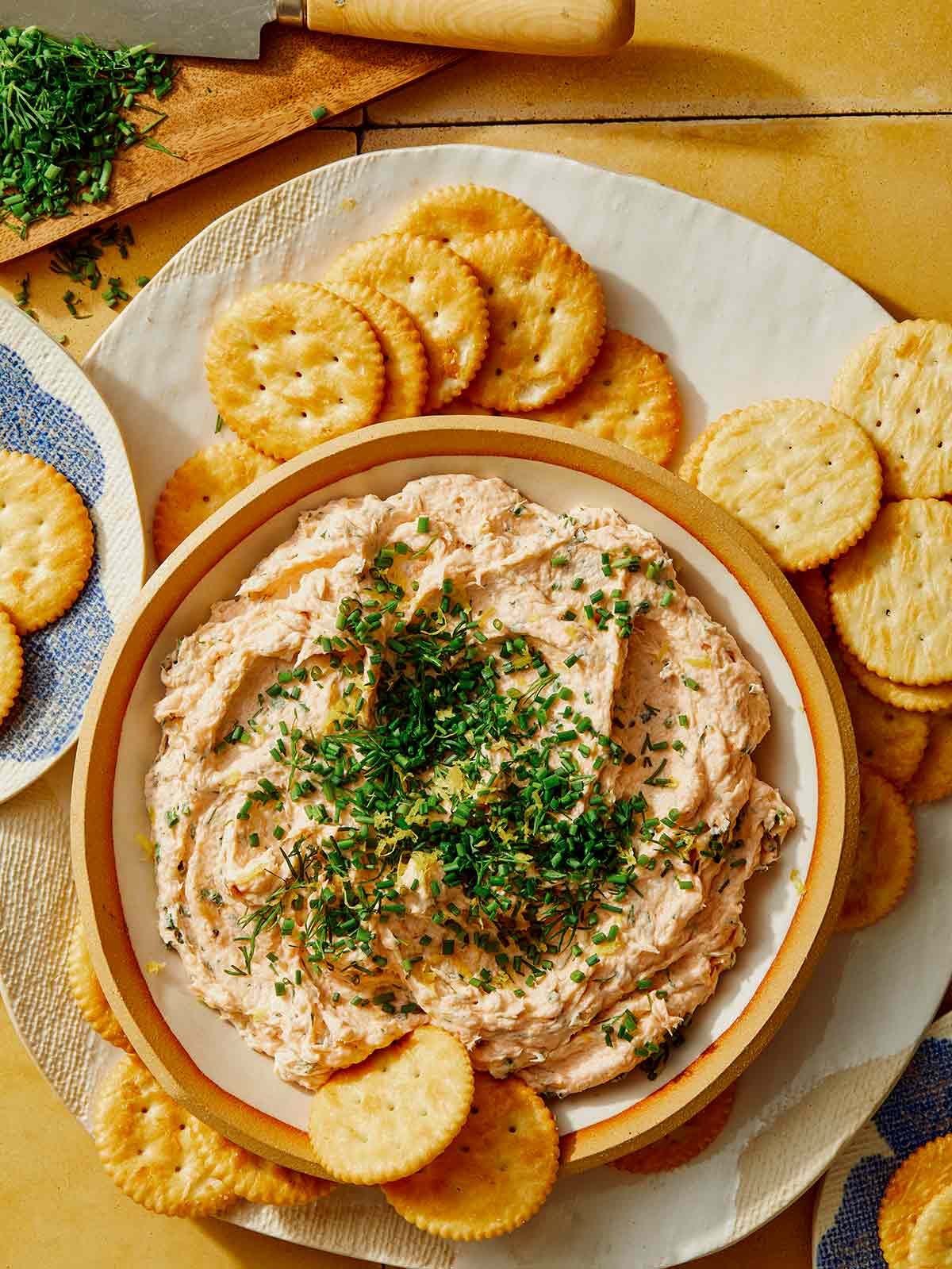 Smoked salmon dip being served with crackers and fresh herbs being chopped on the side.
