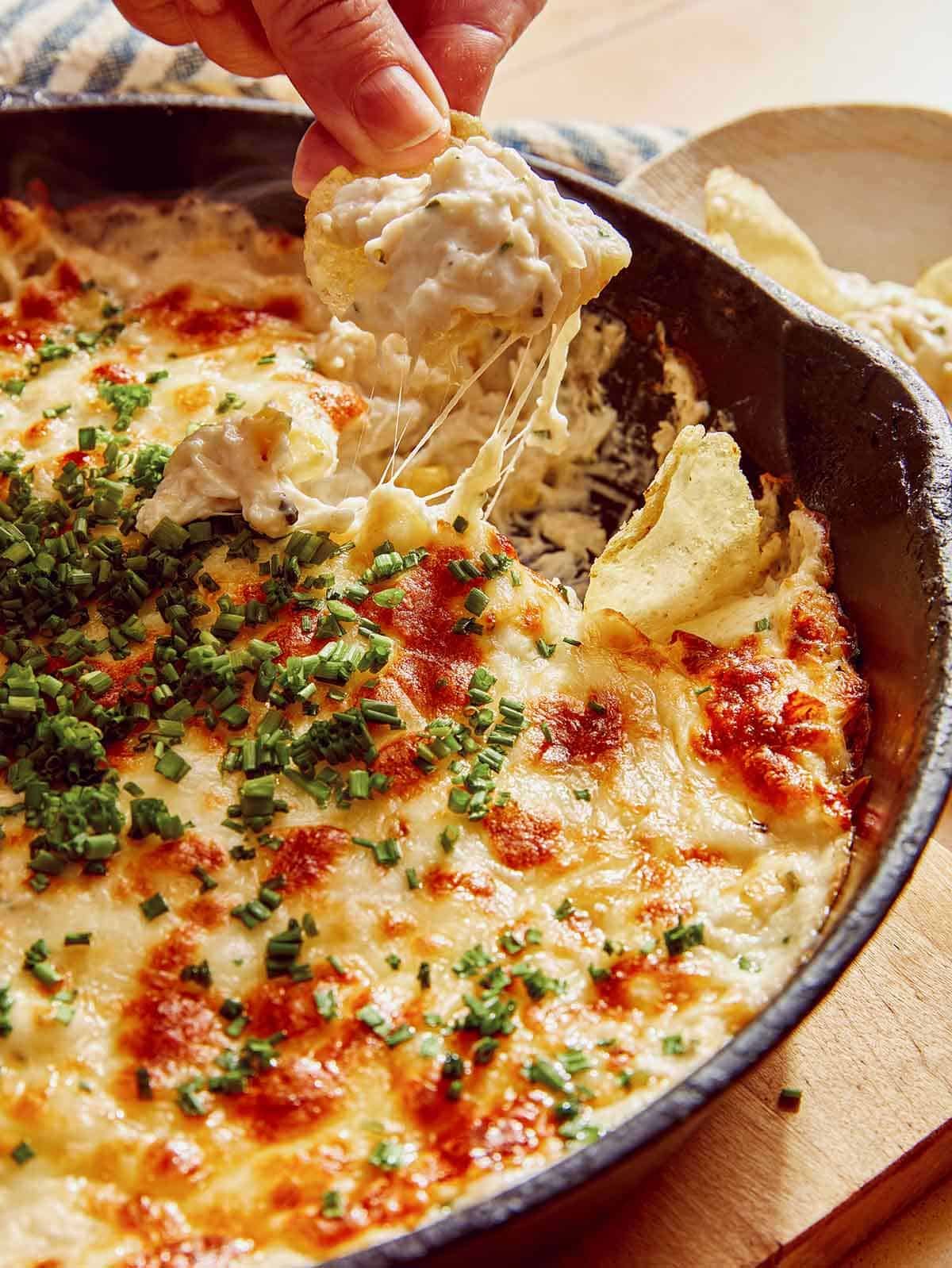 A hand with a potato chip dipping into a skillet of crab dip.
