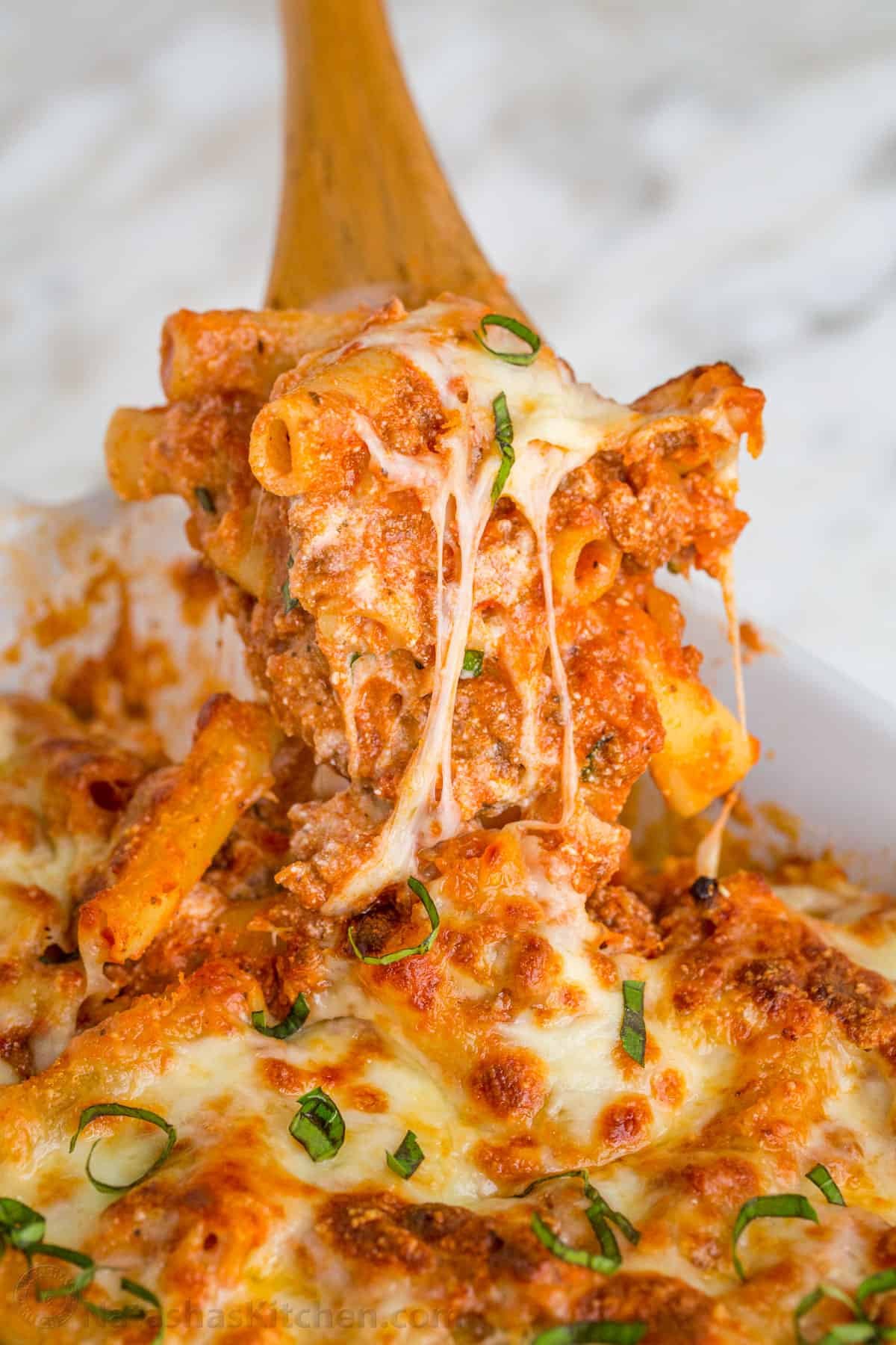 Ground beef pasta casserole Baked ziti being scooped with a wooden spoon, showing the cheese pulls and layers of noodles, meat, cheese, and sauce.