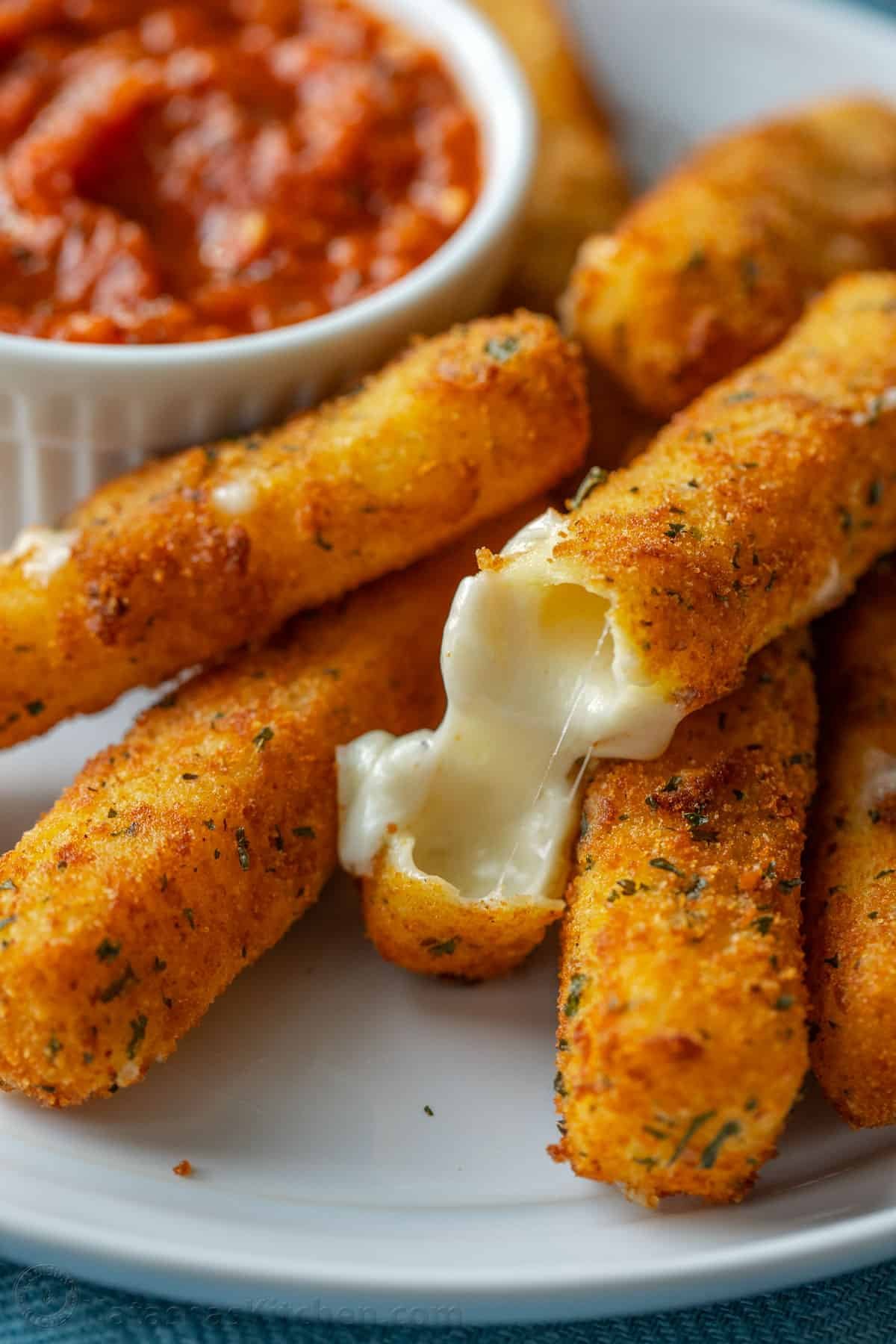 Homemade mozzarella sticks are irresistibly crispy on the outside and gooey and melty in the middle. The ultimate snack or party appetizer! A melty mozzarella cheese stick broken in half in a pile of mozzarella sticks next to a bowl of marinara sauce.