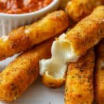 A melty mozzarella cheese stick broken in half in a pile of mozzarella sticks next to a bowl of marinara sauce.
