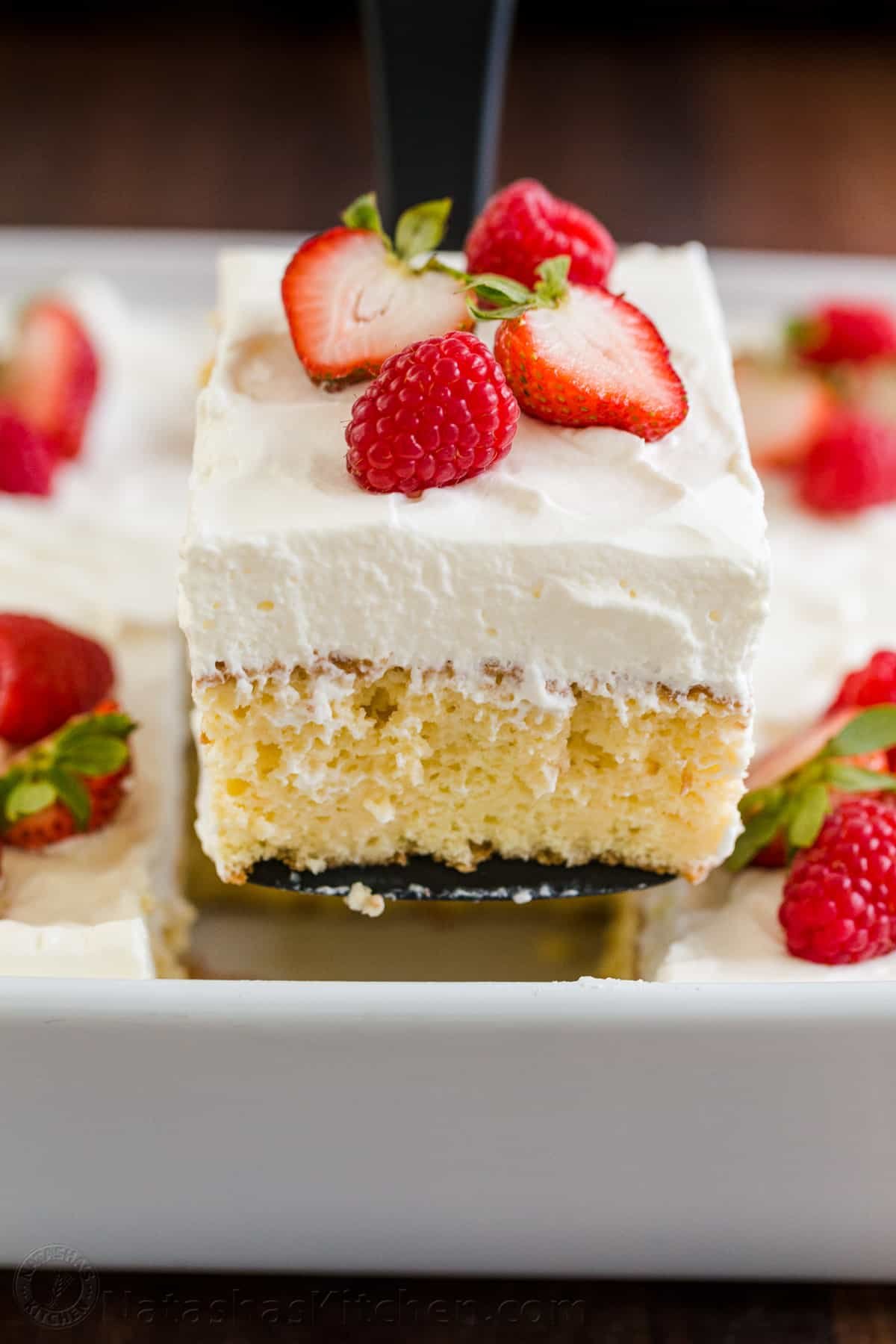 Tres Leches Cake has a soft and ultra-moist crumb. This authentic Tres Leches Cake recipe is soaked with a 3 milk mixture and topped with whipped cream. #tresleches #treslechescake #treslechescakerecipe #mexicancake #cake #cakerecipe #moistcake #natashaskitchen Moist slice of tres leches cake on a spatula with casserole beneath
