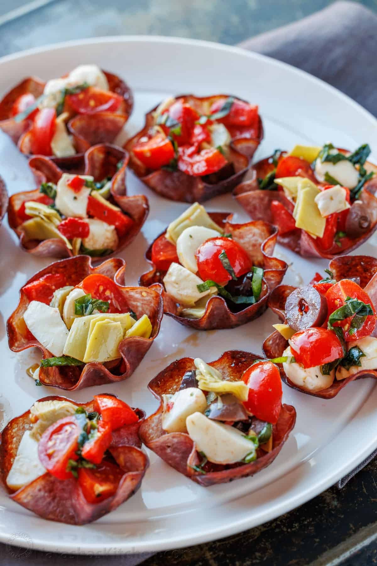 This Salami Cups appetizer recipe is bite-sized perfection! Thin salami baked into crispy cups, filled with your favorite cheeses, antipasto, and herbs. Salami Antipasto Bites appetizer displayed on a serving platter