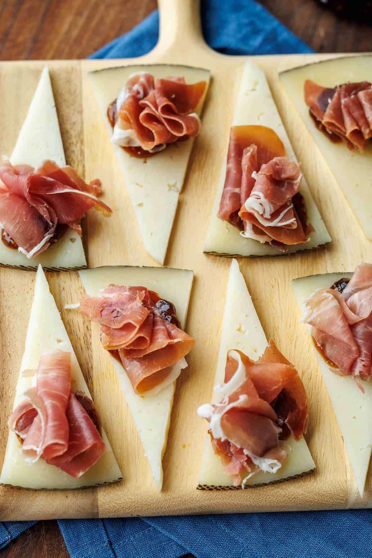 Easy manchego cheese bites with fig preserves and prosciutto take less than 5 minutes to make, but they look elegant and taste divine! Manchego Cheese Bites with fig jam and prosciutto on a wooden cutting board