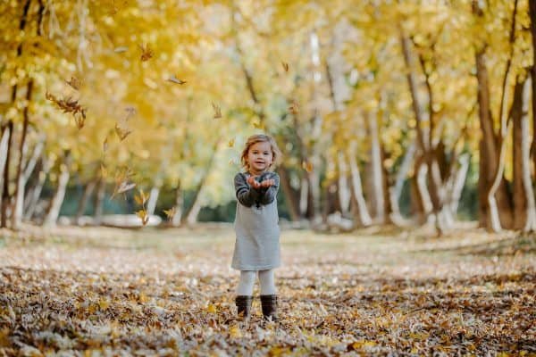 A little girl that is standing and playing in the leaves 