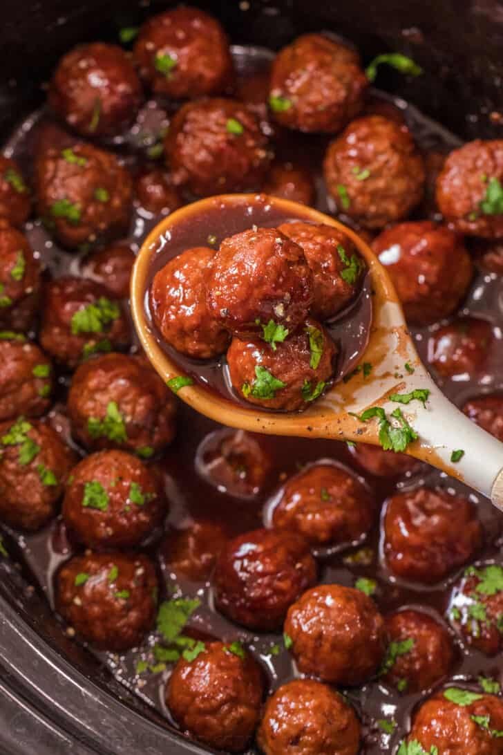 Tender and juicy grape jelly meatballs smothered in a sweet and spicy sauce and slow-cooked to perfection. A ladle full of grape jelly meatballs topped with chopped greens.
