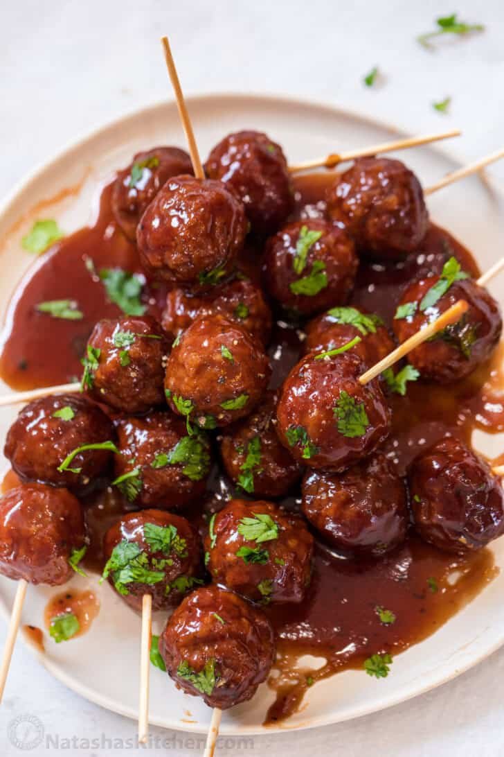 The ultimate party appetizer recipe, these grape jelly meatballs are juicy and packed with flavors slow cooked in a sweet and sour sauce. A white plate loaded with meatballs topped with chopped greens.