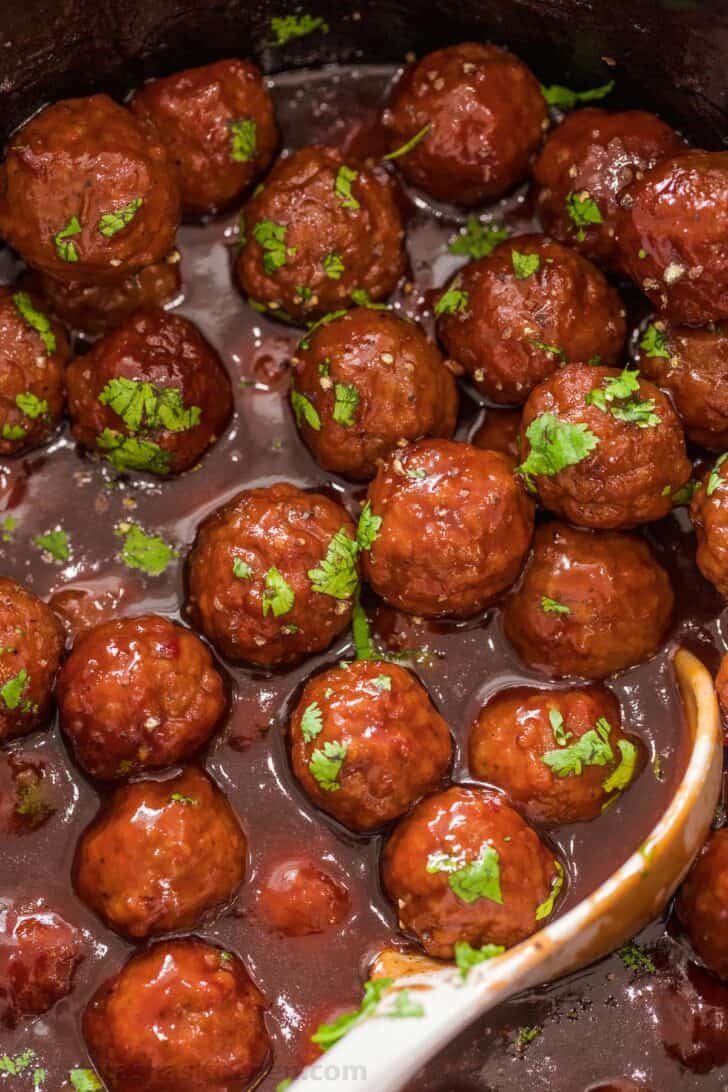 The BEST homemade grape jelly meatballs that give you the juicest, tender meatballs smothered in a sweet and sour sauce. Grape jelly meatballs in a slow cooker topped with fresh chopped greens with a ladle.