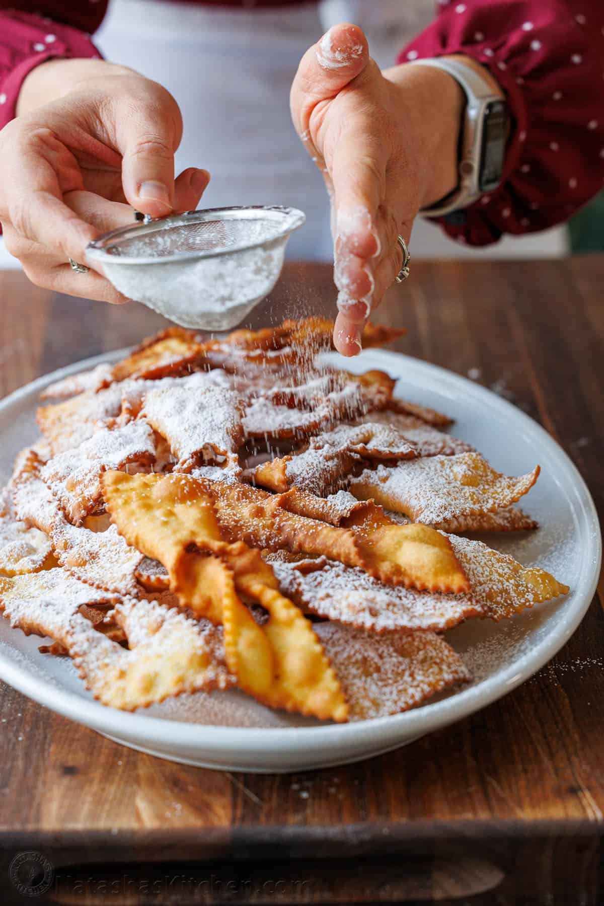 Traditional Angel Wing Cookies (Polish Chrusciki) make a beautiful holiday cookie. These ribbon cookies are dusted with sugar & irresistible. Dusting Angel Wing Cookies Polish Chrusciki with powdered sugar on a white platter