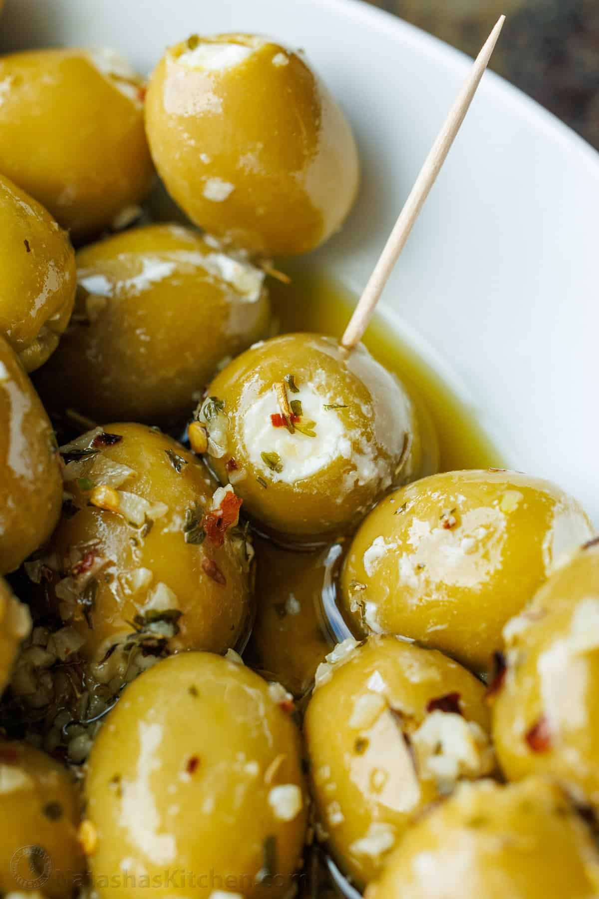 Stuffed Olives are the ultimate no-bake appetizer—quick to make, yet fancy enough for any occasion. They're filled with garlic and herb Boursin cheese and marinated in a flavorful garlic and herb olive oil. A close-up of stuffed olives in marinade, in a white bowl served on toothpicks.