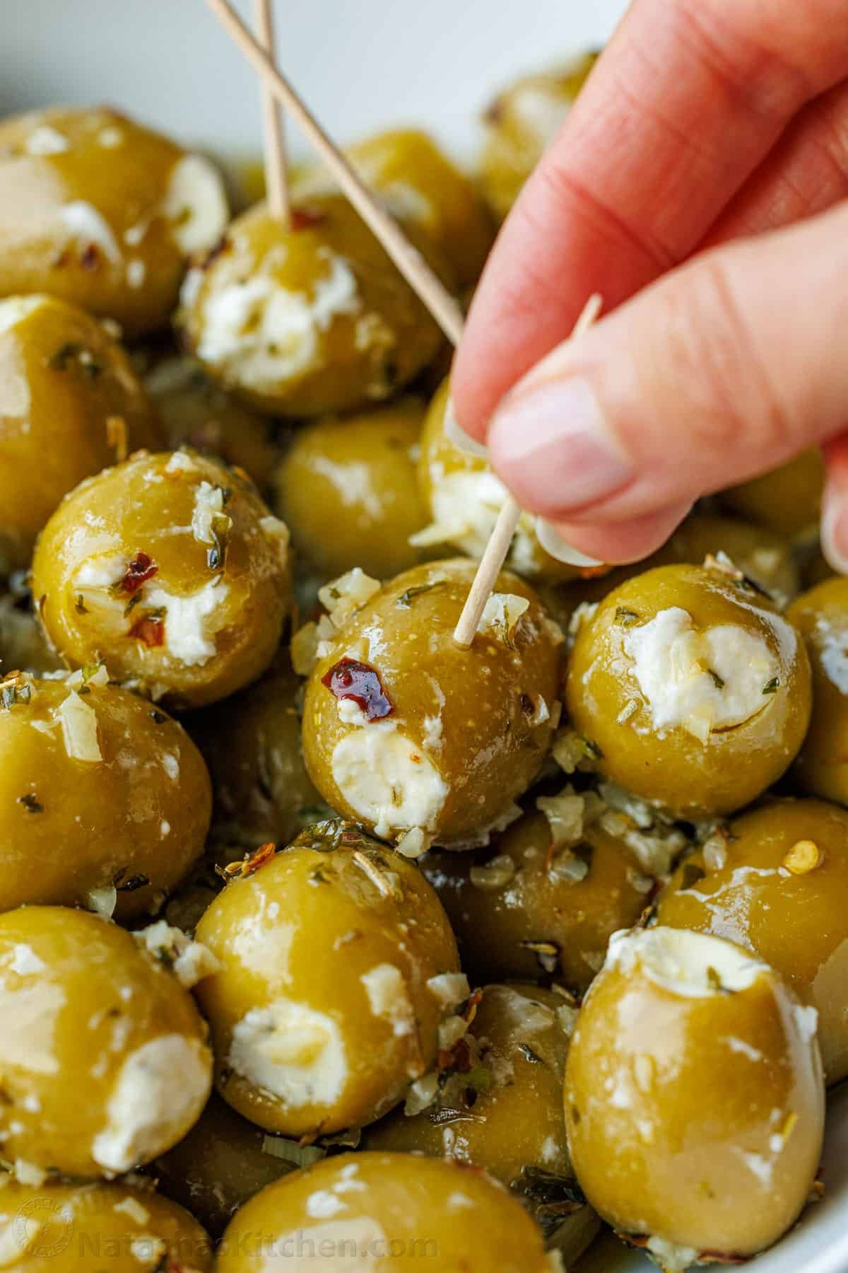 Stuffed Olives are the ultimate no-bake appetizer—quick to make, yet fancy enough for any occasion. They're filled with garlic and herb Boursin cheese and marinated in a flavorful garlic and herb olive oil. A close-up of stuffed olives served on toothpicks, with a hand delicately pinching one toothpick to pick up an olive from the batch.