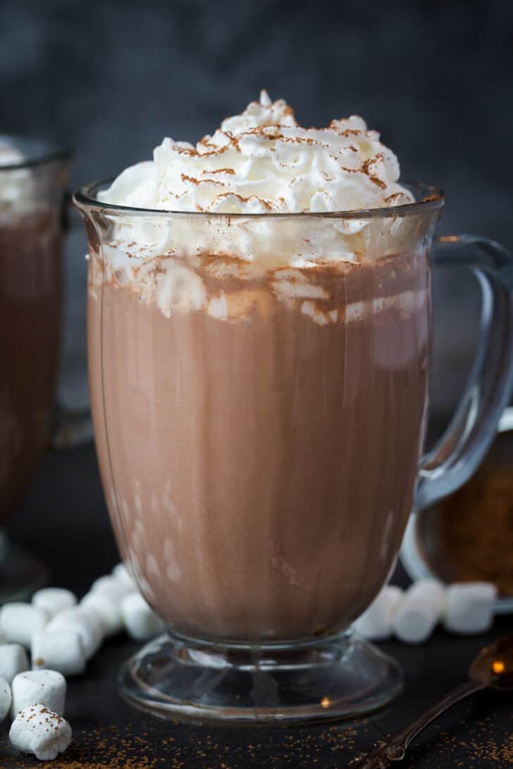 Simple recipe for the creamiest hot chocoalte that is rich in flavor and ready in under 30 minutes! A close up of hot chocolate in a glass mug with whipped cream and cocoa powder.