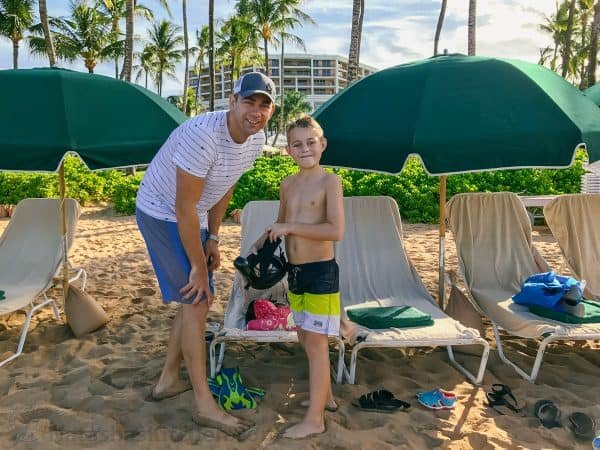 A father and son smiling at the camera, standing next to beach chairs and umbrellas 
